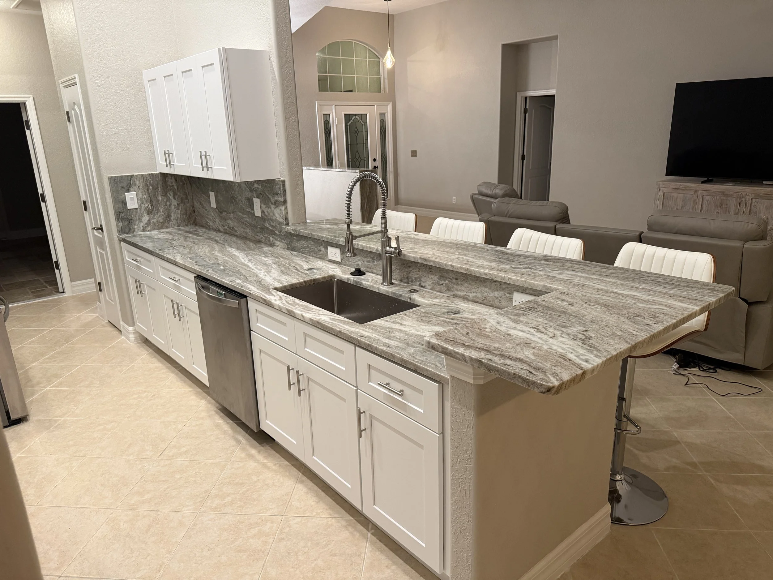 Modern kitchen with granite countertops, white cabinets, stainless steel dishwasher, and a large kitchen island with barstools. Open-concept living space with beige walls, a flat-screen TV, and beige couches.
