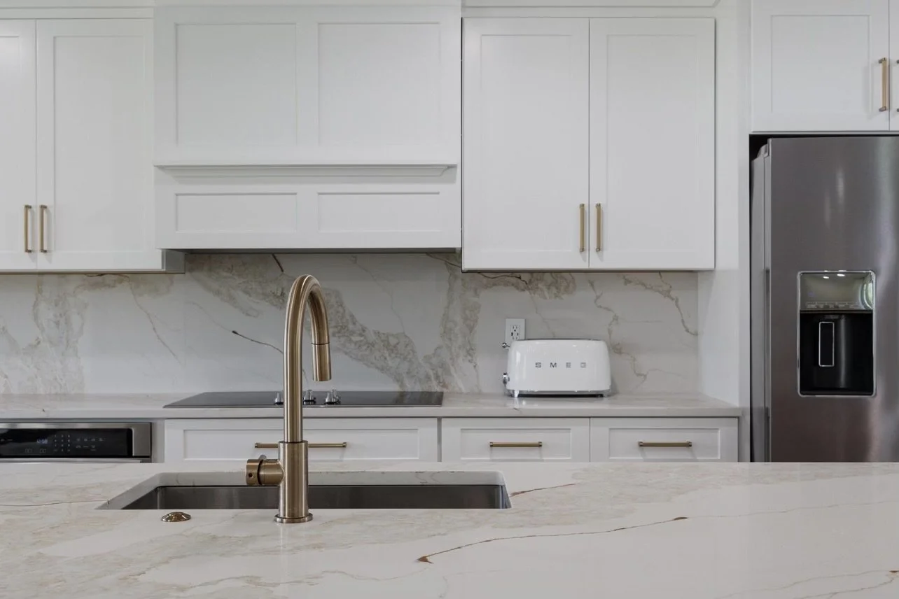 Modern kitchen with white cabinets, a marble backsplash, stainless steel refrigerator, a white toaster on the counter, and a gold faucet over a double sink.