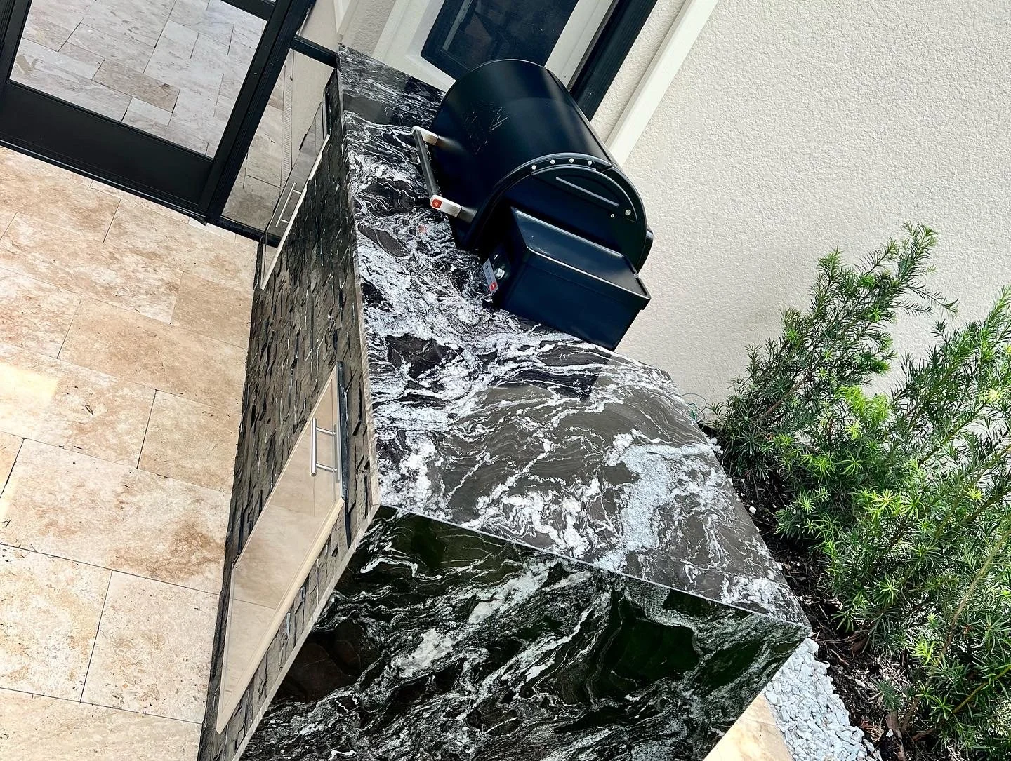 Black marble countertop with a built-in stainless steel sink, a black portable grill, and a small grill box outdoors near a door with tiled flooring and a plant to the right.