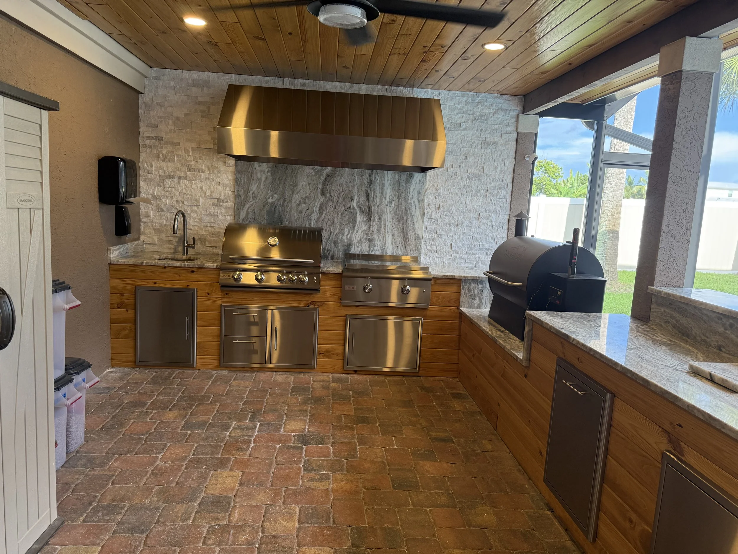 An outdoor kitchen area featuring stainless steel appliances, a wooden ceiling, stone wall, and brick flooring, with a large window showing a backyard with green grass and trees.