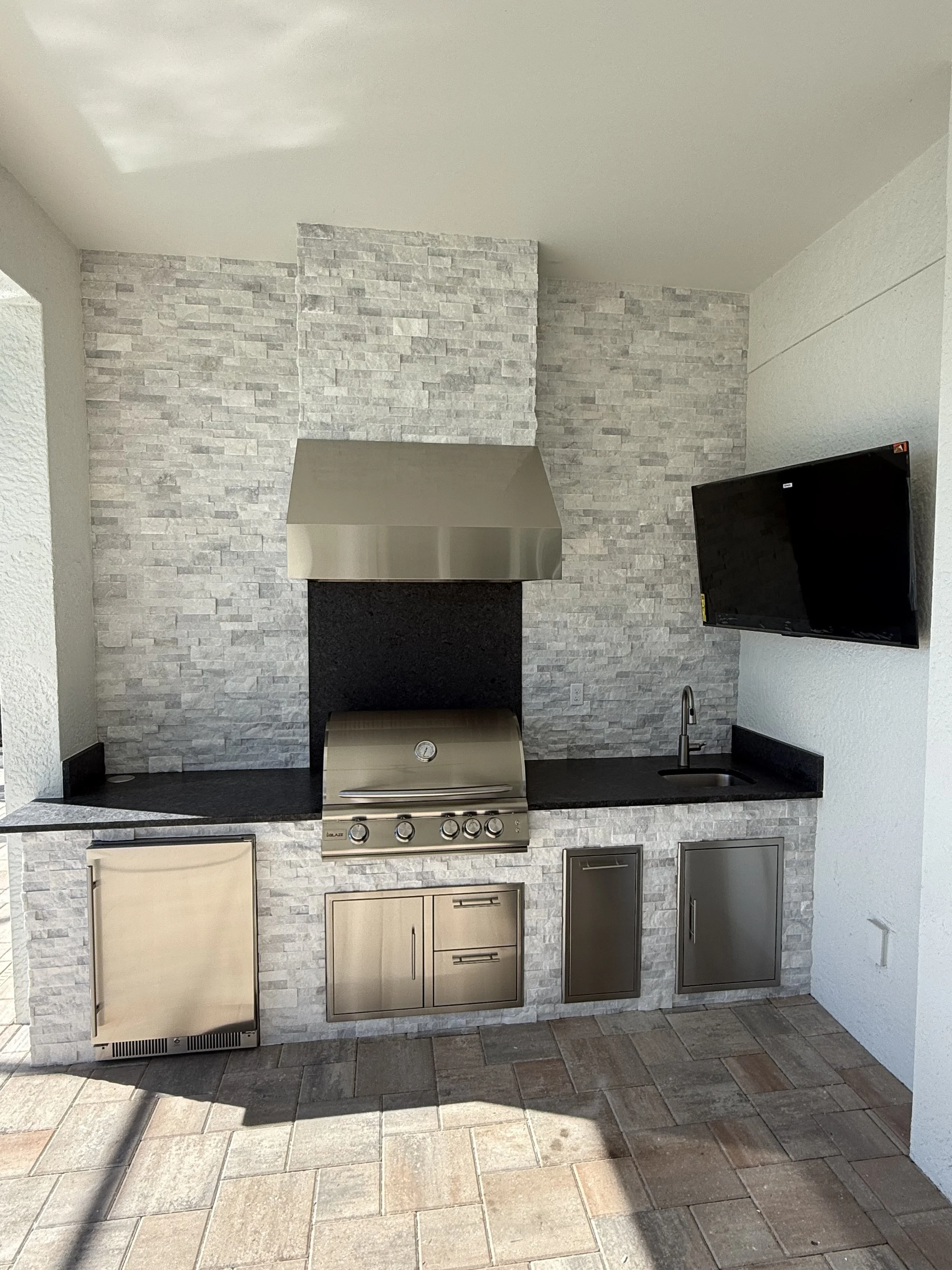 Outdoor kitchen with a countertop, stainless steel grill, and small sink set against a textured stone wall.