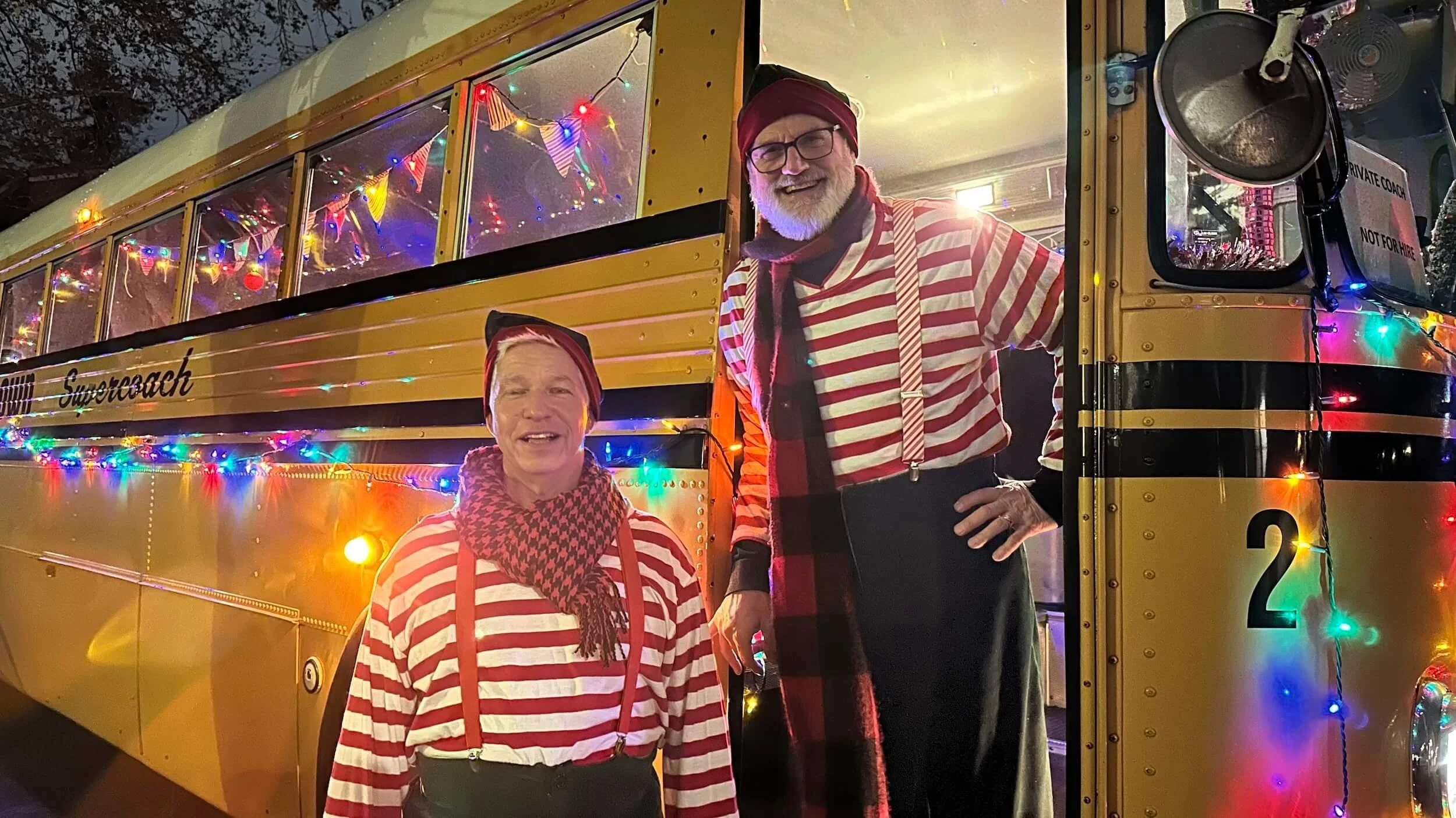 Two people dressed in striped shirts and scarves, smiling in front of a decorated yellow bus with colorful string lights, at night.