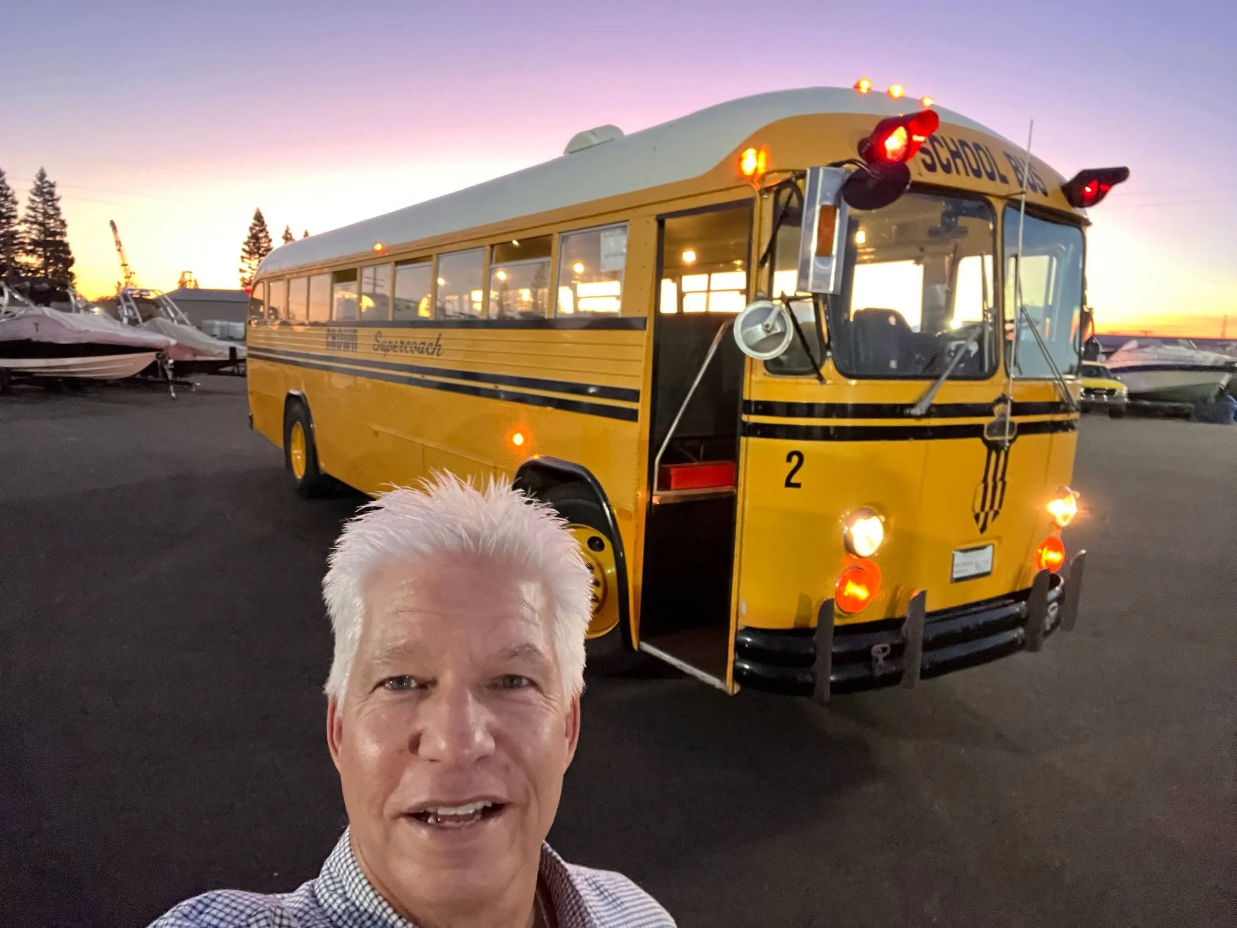 A person with white hair taking a selfie in front of a yellow school bus with flashing red lights. The background features a school bus during sunset or sunrise with a clear sky and some trees.