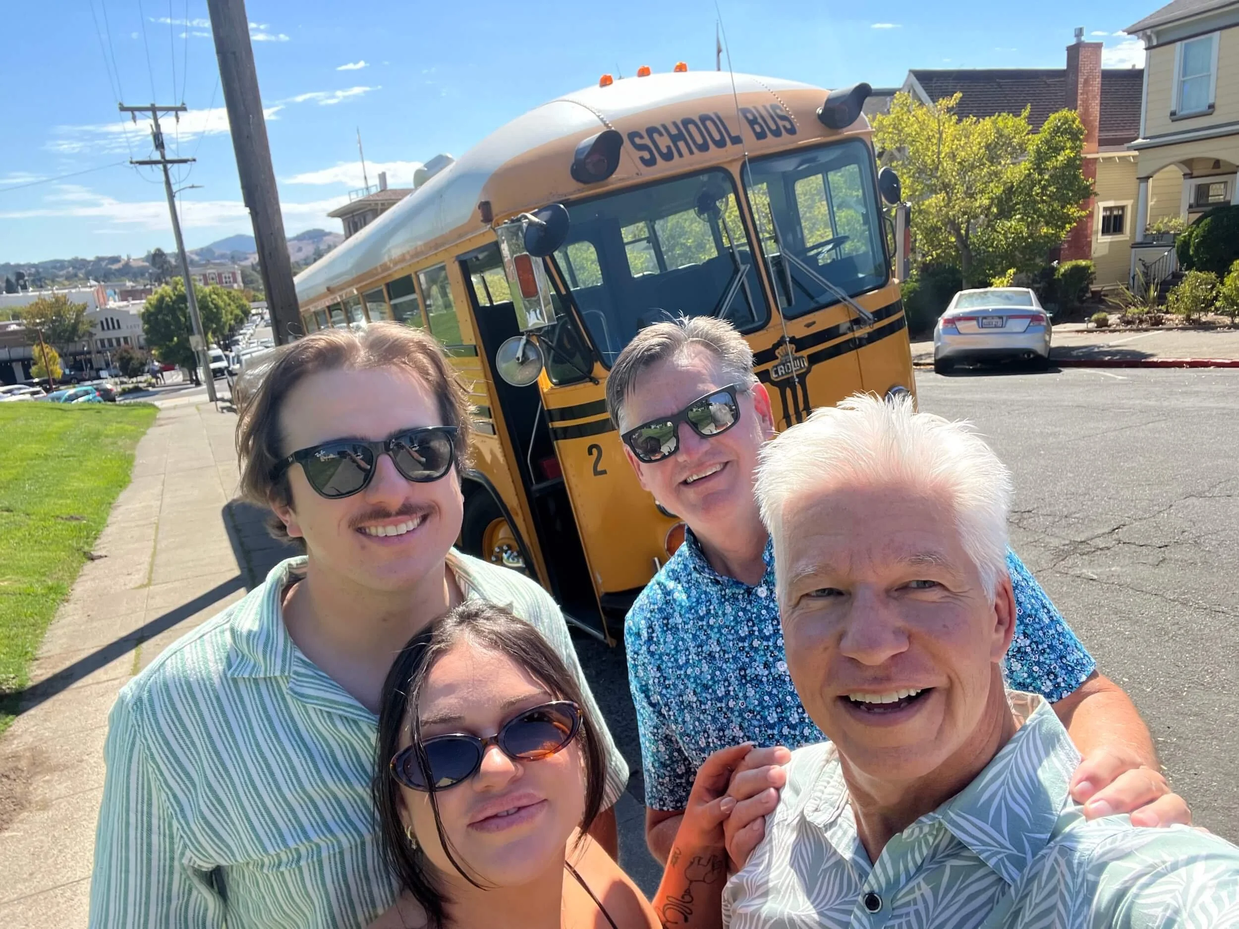 A group of people taking a selfie outside on a sunny day, with a school bus and houses in the background.