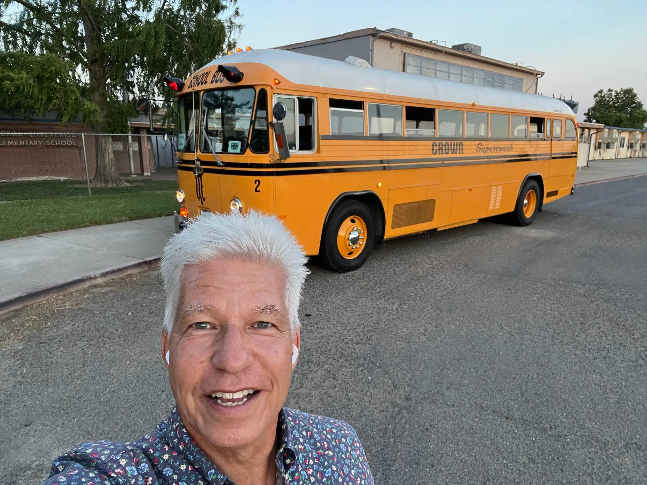 A man with gray hair smiling for a selfie in front of a yellow school bus labeled 'CROWN Supercoach' parked on a street, with a fence and trees in the background.