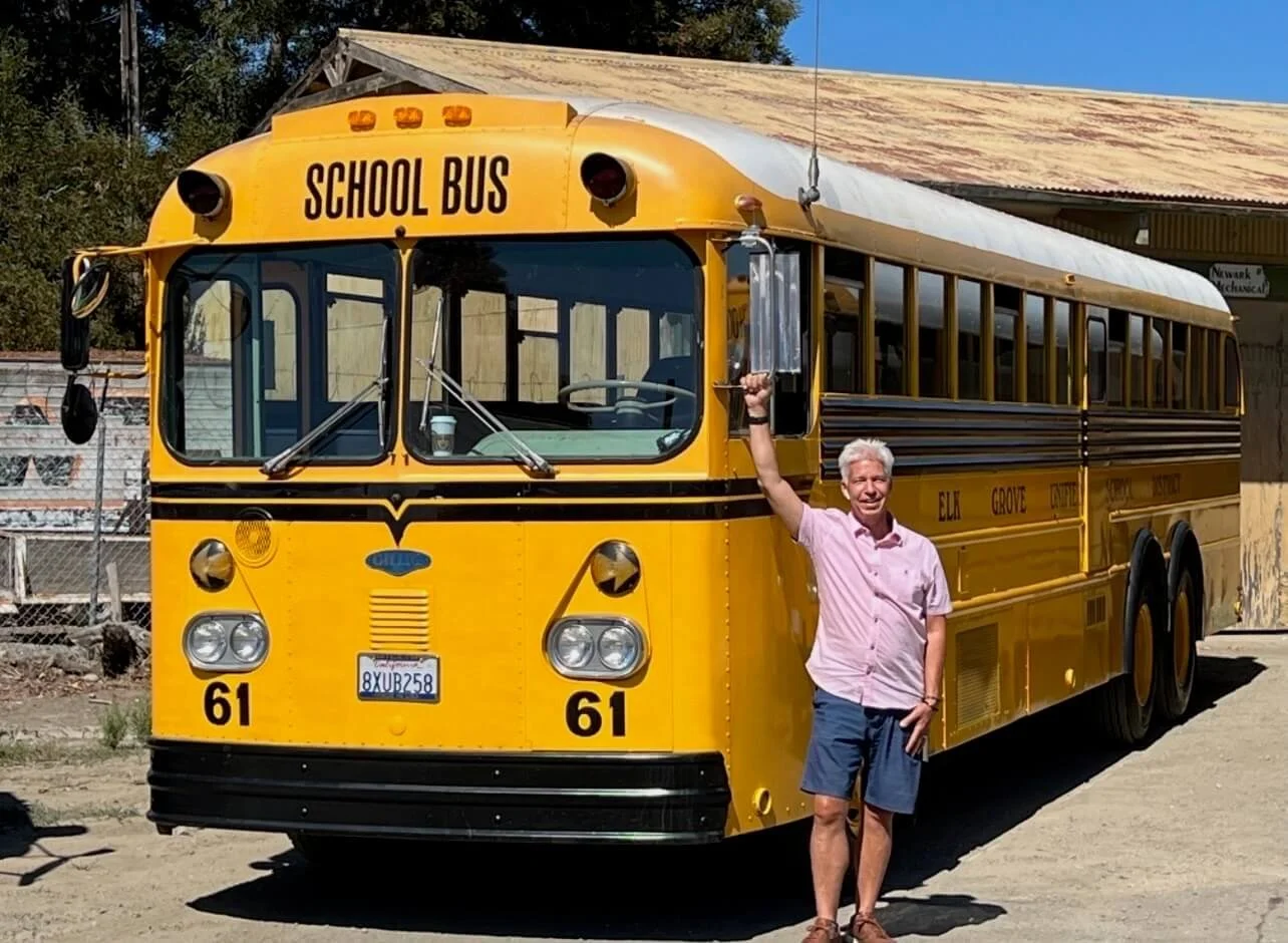 A man in a pink shirt and shorts standing next to a vintage yellow school bus, raising his arm and holding a small object. The bus has the words 'School Bus' on the front and the number 61, with text indicating it is from Elk Grove.