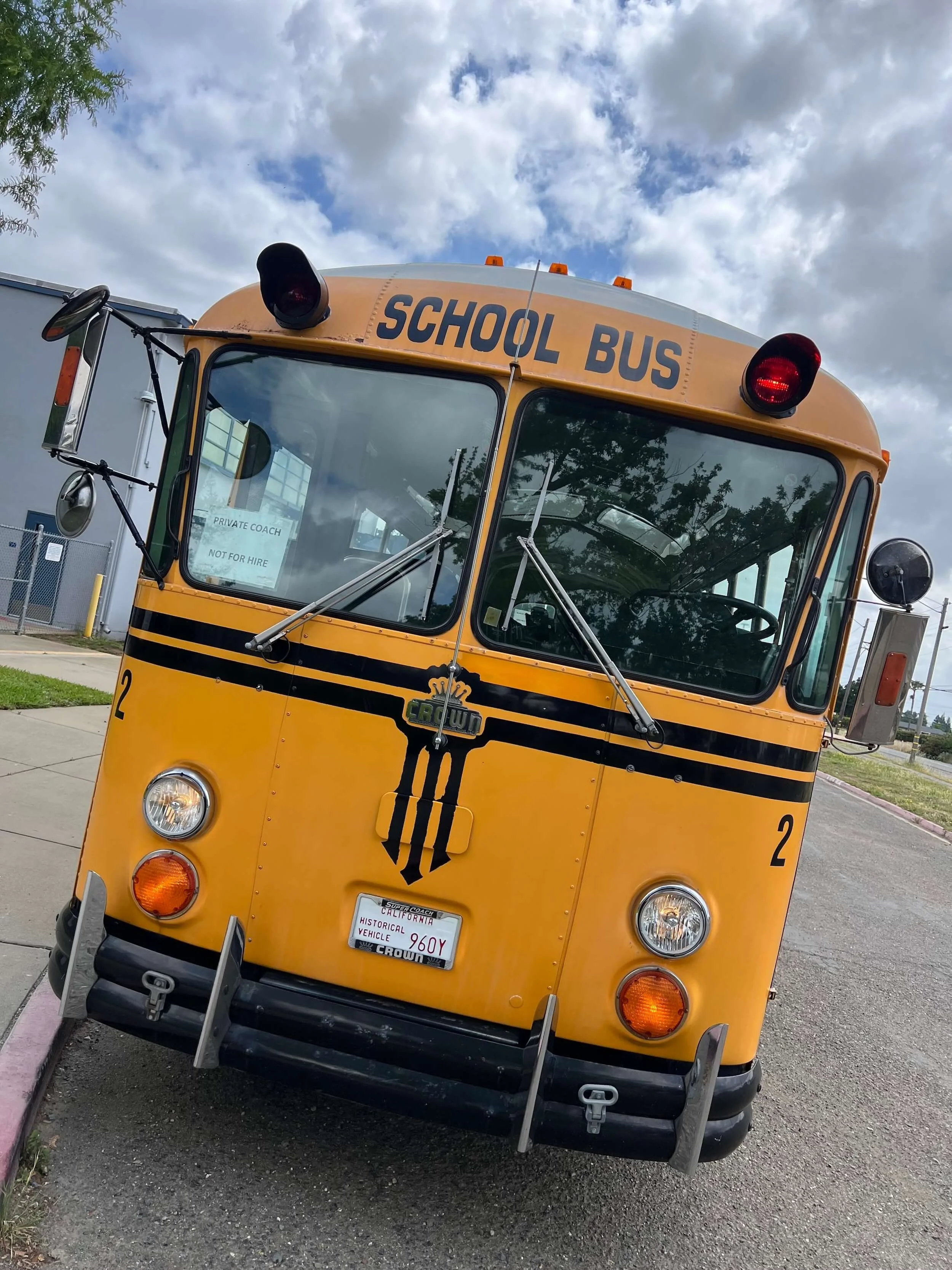 Front view of a vintage yellow school bus with black stripes, parked on the side of a street, with a sign in the windshield that reads 'Private Coach Not For Hire' and a California license plate The bus has round headlights and orange warning lights on the roof, with a blue sky and some clouds in the background.