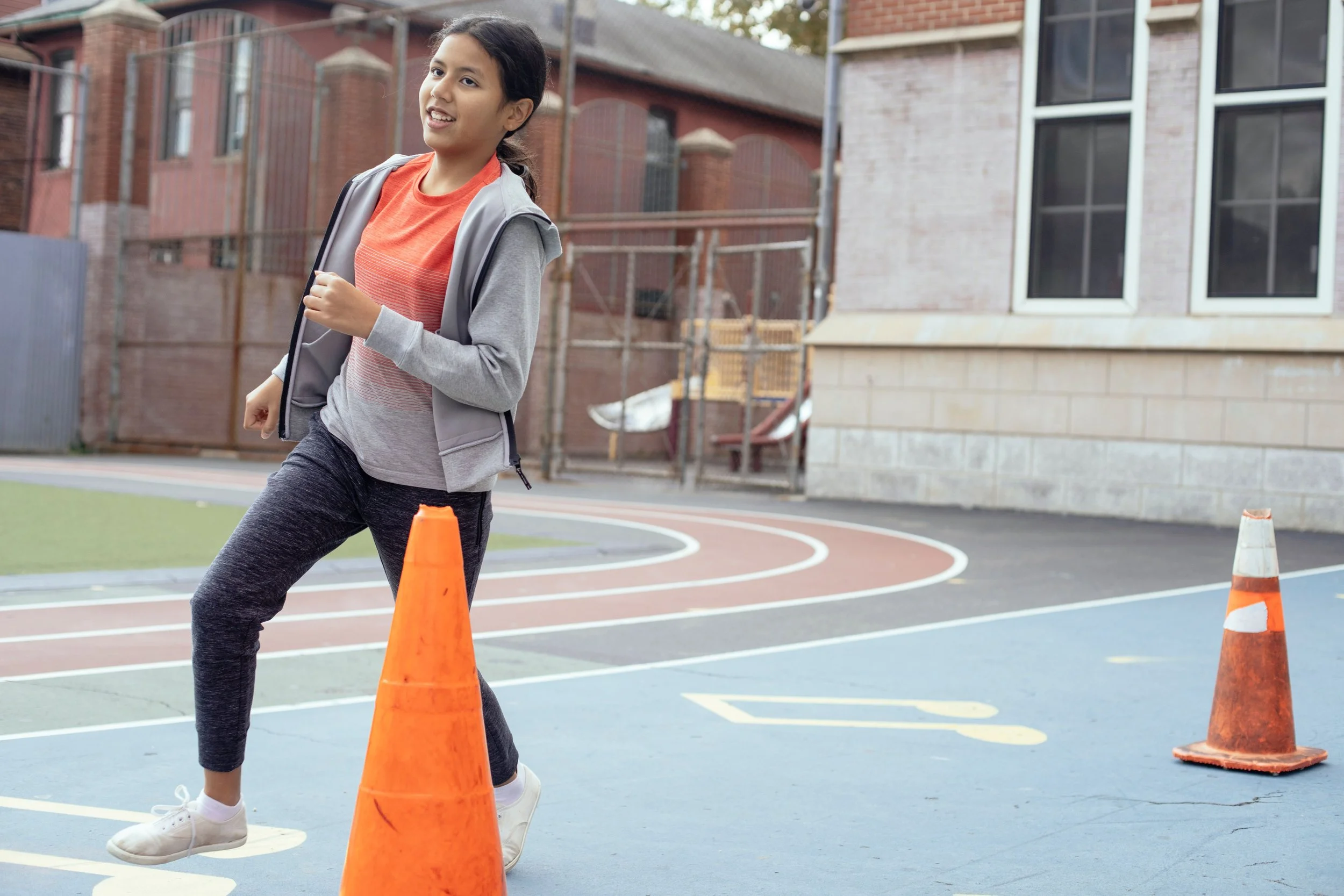 A young girl with dark hair in a ponytail jogging between orange traffic cones on a sports court outside a brick building.