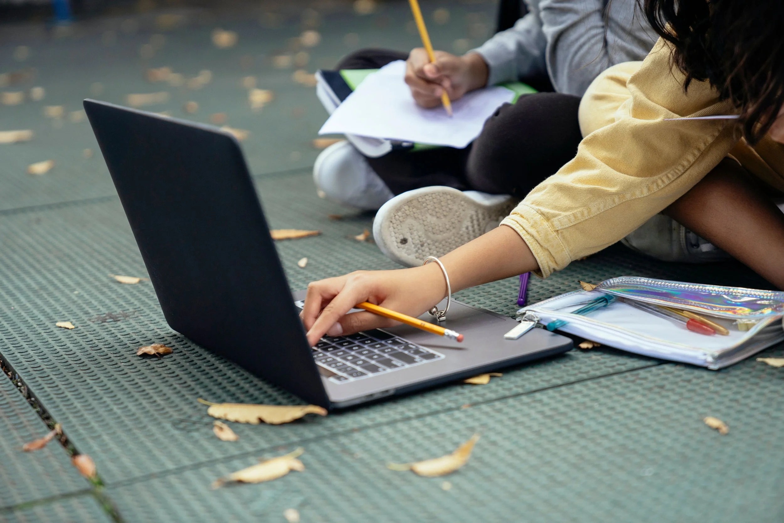 People sitting on a green outdoor bench working on a laptop, with notebooks and pens. Fallen leaves surround them.