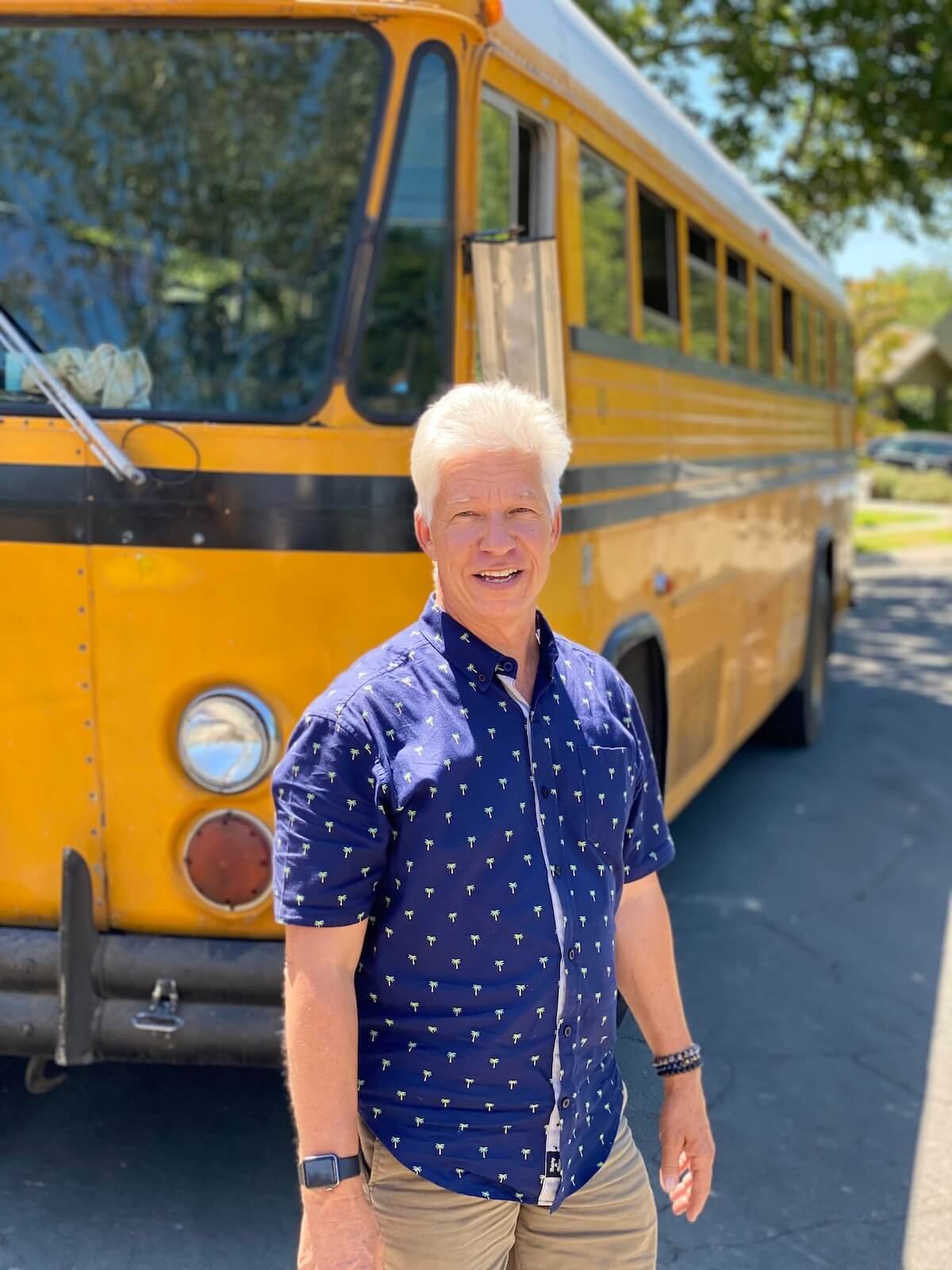 An older man with white hair smiling, wearing a blue shirt with small palm tree patterns and khaki shorts, standing in front of a yellow school bus on a sunny day with green trees in the background.