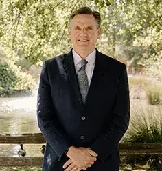 A man in a dark suit standing outdoors near a body of water, smiling with trees in the background.