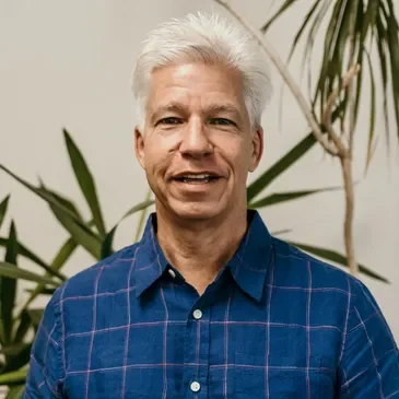 A middle-aged man with white hair is smiling in front of green plants, wearing a blue checkered shirt.