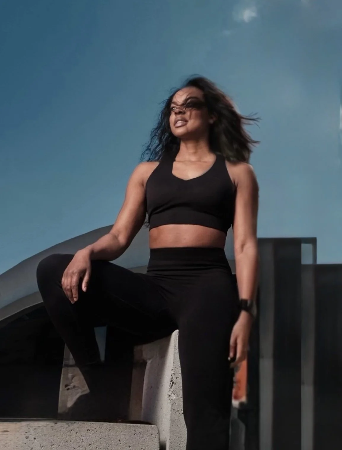 A woman in black athletic wear posing outdoors on a concrete surface against a blue sky.