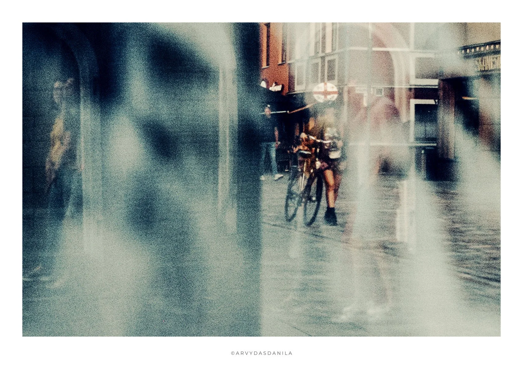 A person holding a bicycle on a rainy city street with reflections and water spray visible.