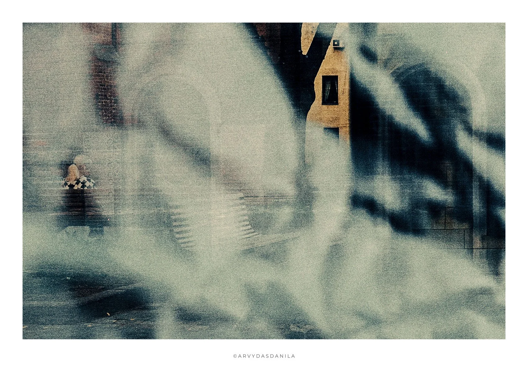 A blurry, abstract photograph featuring a person with blonde hair and checkered clothing walking in a rain-soaked, urban environment with reflections and a building in the background.