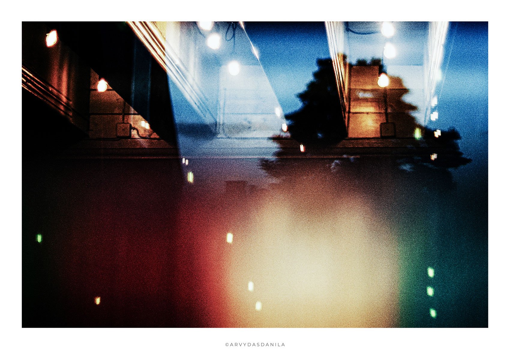 Reflection of a building and trees with string lights on a glass surface, creating a colorful, abstract scene at dusk or night.