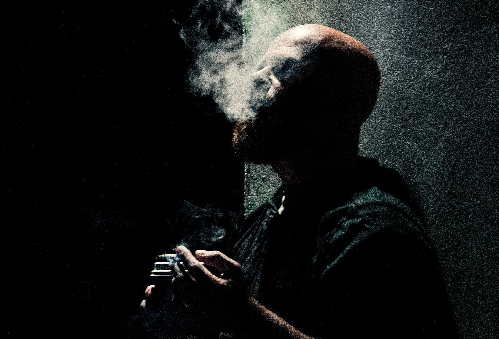 A person with a bald head and beard standing against a textured wall, holding a smartphone, with smoke or vapor coming from their mouth.