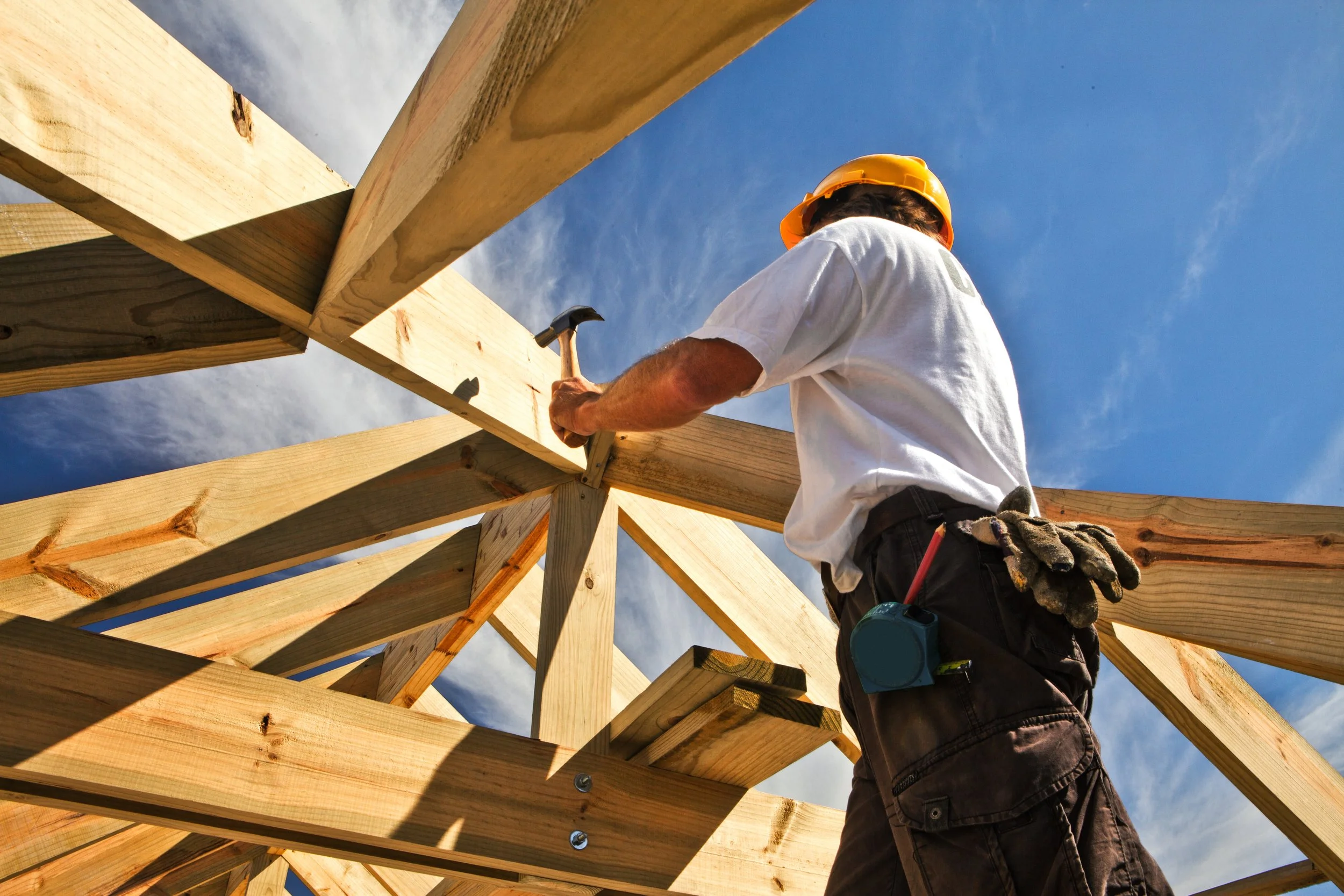A construction worker wearing a yellow safety helmet and white T-shirt, hammering a nail into wooden beams during daylight under a blue sky with few clouds.