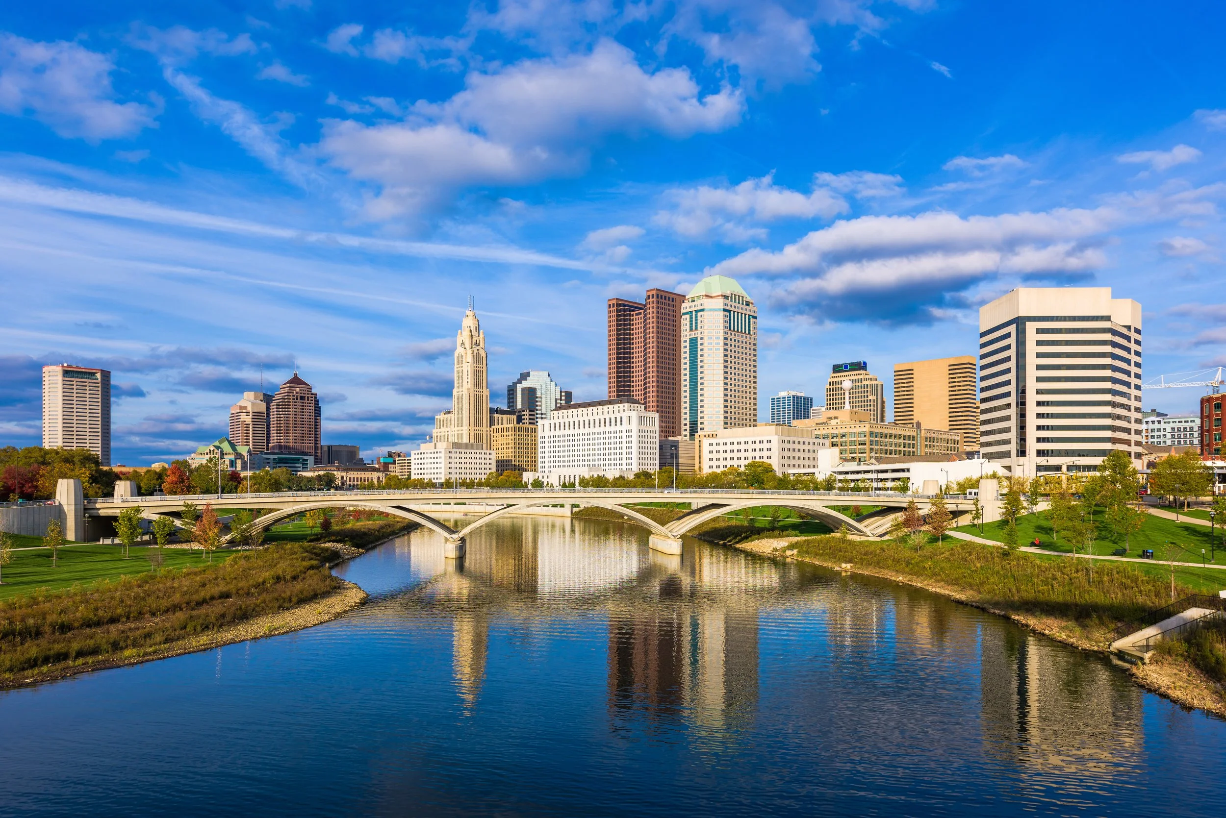 A city skyline with tall buildings reflected in a river, under a bright blue sky with scattered clouds, with a bridge crossing the river in the foreground.