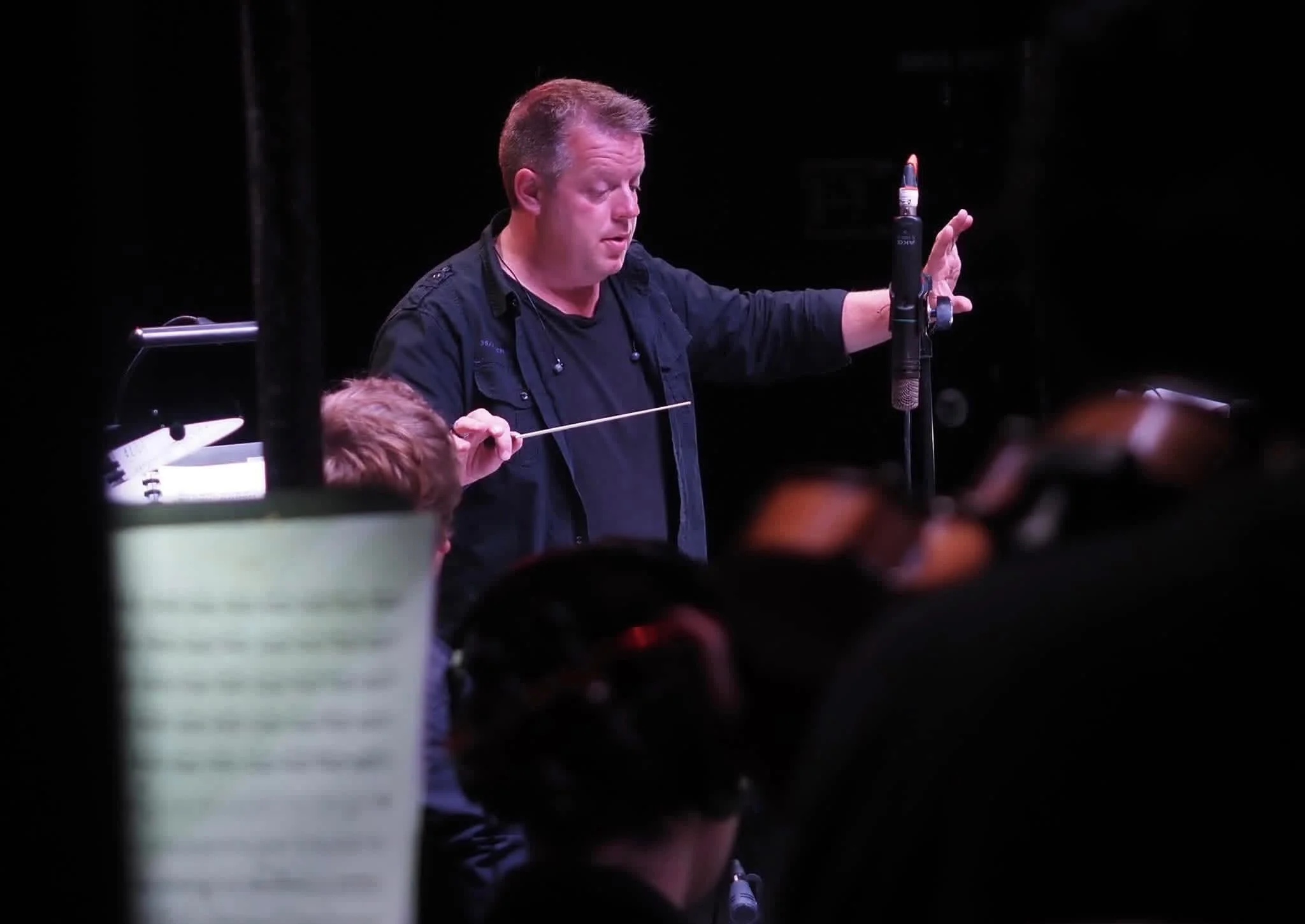 A man with short hair, dressed in black, standing on a stage with a dark background, with his eyes closed, and holding an object in his right hand while resting his left hand on a microphone stand.