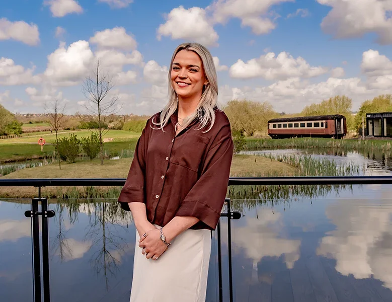 A woman with blonde hair smiling stands outdoors on a balcony over a pond with trees and a vintage train in the background, under a partly cloudy sky.