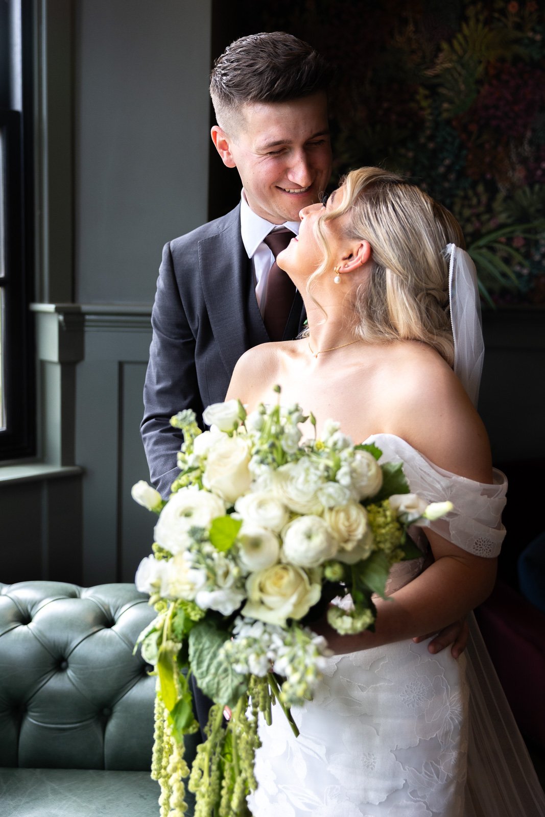 A newlywed couple sharing a joyful moment indoors. The groom is dressed in a gray suit with a tie, and the bride is in a white wedding gown holding a large bouquet of white flowers. They are smiling and looking at each other affectionately.