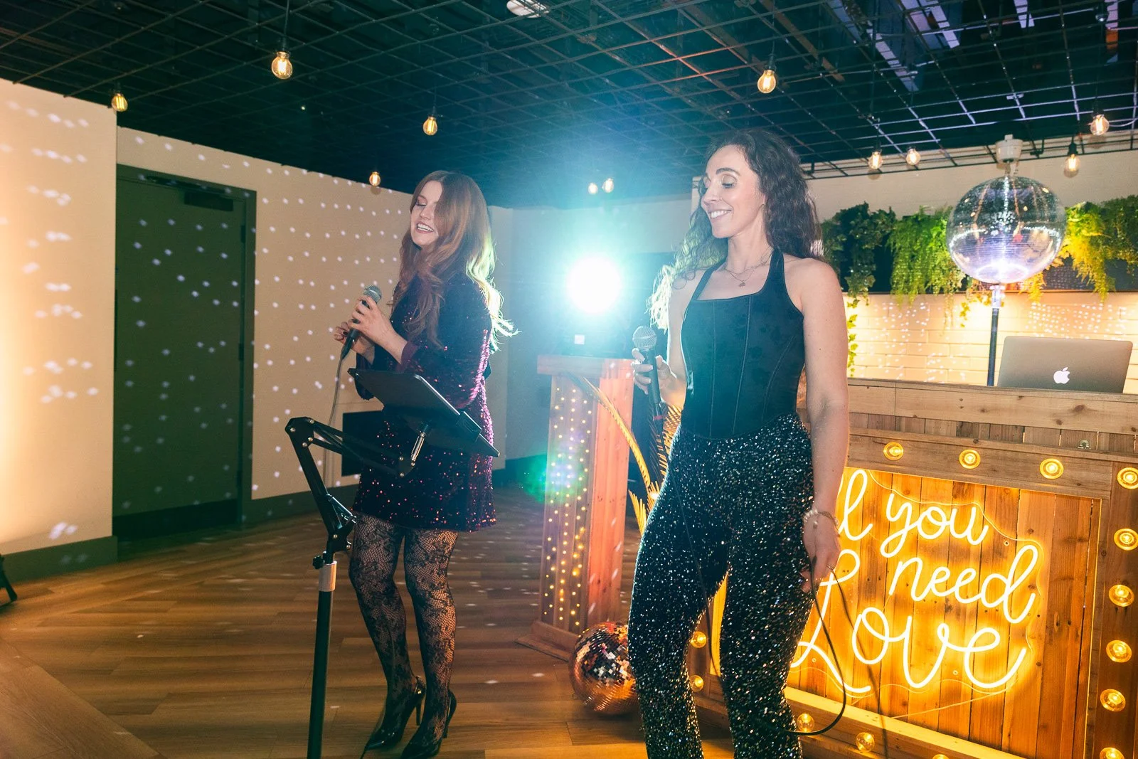 Velvet Harmony with microphones at a lively indoor party or celebration, decorated with string lights, a glowing sign that says 'All you need is love', and a disco ball.