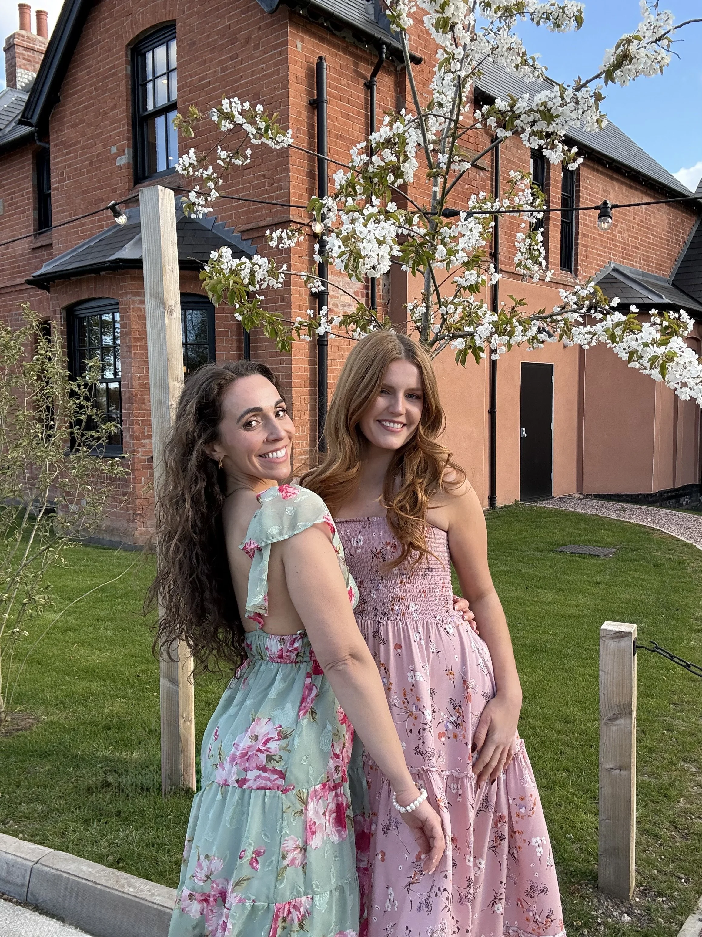 Velvet Harmony in floral dresses standing outdoors on a grass lawn, smiling at the camera, with a flowering tree, wooden fence posts, and a brick house in the background.