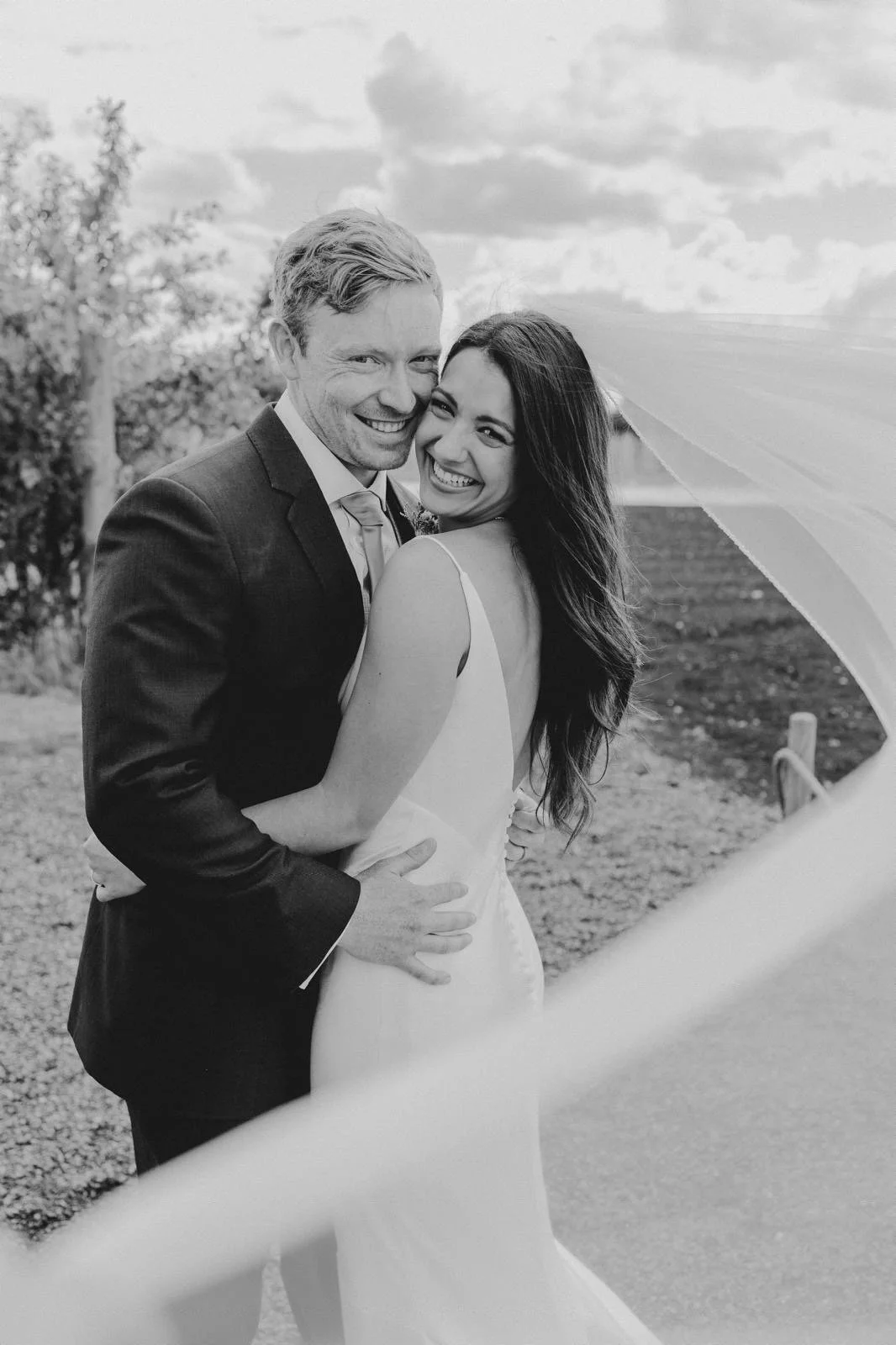Black and white photo of a smiling newlywed couple embracing outdoors, with a tree and cloudy sky in the background.