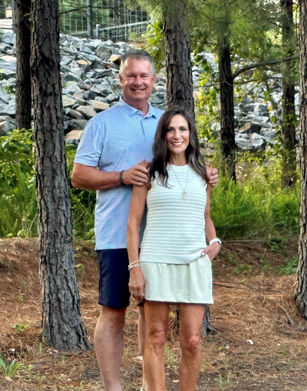 A smiling couple standing outdoors in a wooded area with tall trees and rocks in the background. The man has short gray hair, wears a light blue shirt and dark shorts, and stands behind the woman with one hand on her shoulder. The woman has long dark hair, wears a sleeveless striped top and a skirt, and has her hands resting in her pockets.