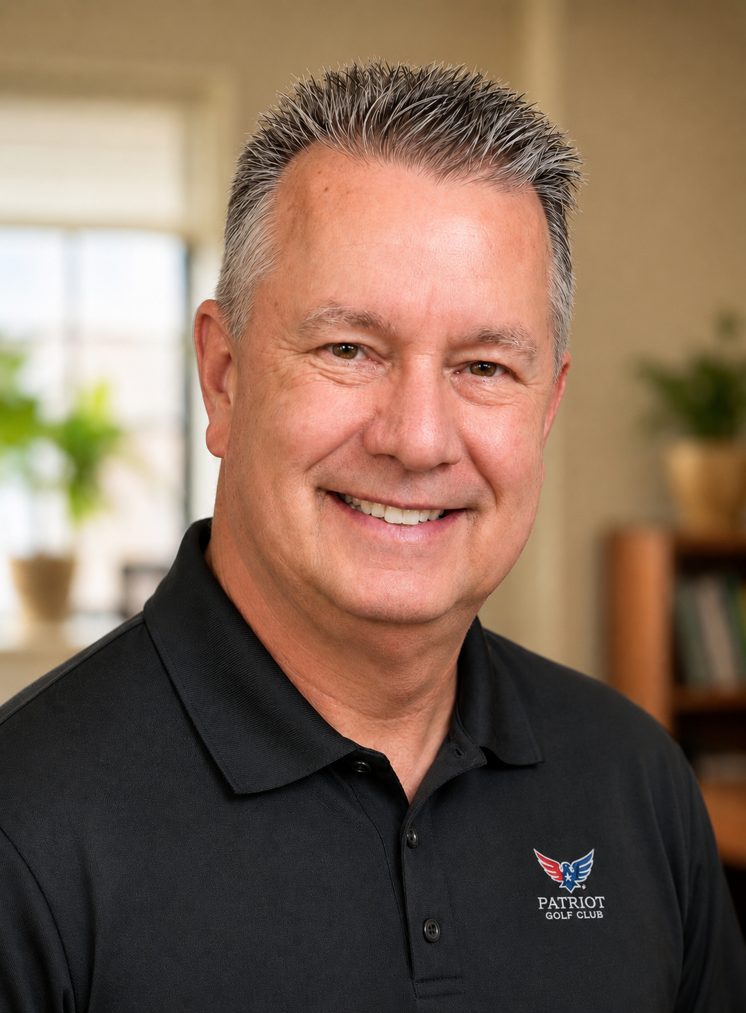 A middle-aged man smiling, wearing a black polo shirt with a Patriot Golf Club logo, in a room with a window, plants, and bookshelves in the background.