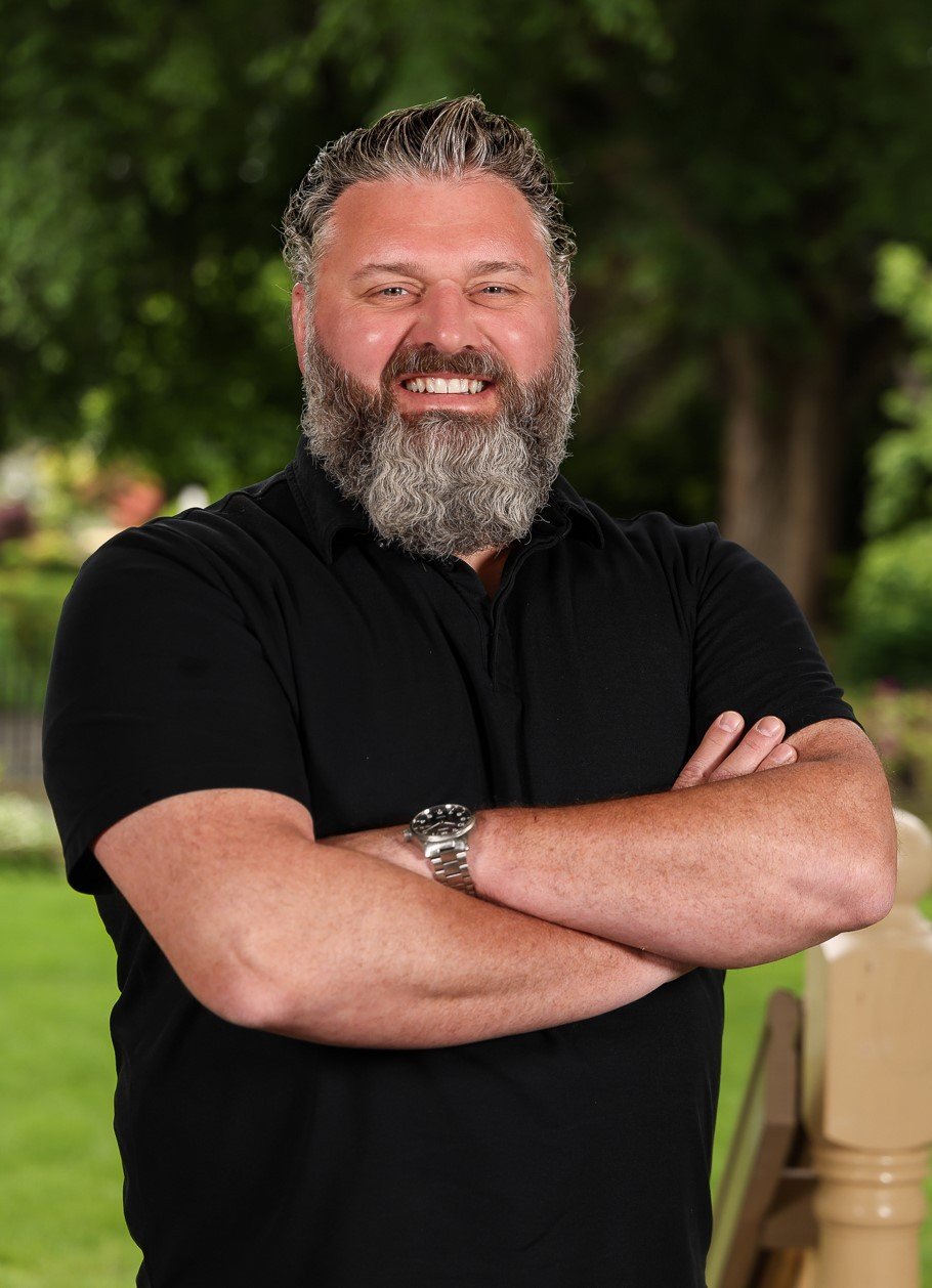 A man with a gray beard smiling, wearing a black shirt and a silver watch, standing outdoors with green trees in the background. Local mortgage professional in Washington State who lives and works in Bellingham, WA.