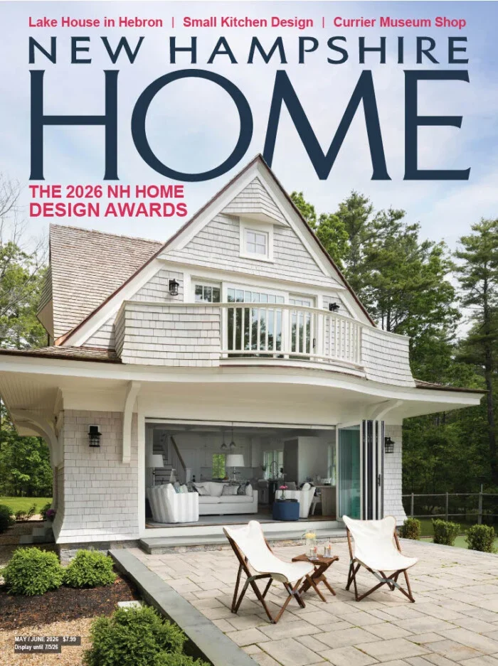 Cover of New Hampshire Home magazine featuring a two-story white house with a small upper balcony, large living room with white furniture visible through open glass doors, and two chairs with a small table outside on a stone patio. The magazine highlights the 2026 NH Home Design Awards.