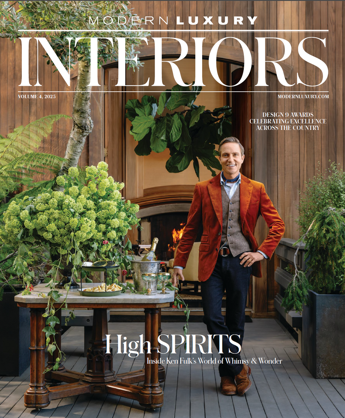 Cover of Modern Luxury Interiors magazine featuring a man standing in a cozy, wood-paneled room with fireplace, surrounded by lush green plants and a table with drinks and snacks.