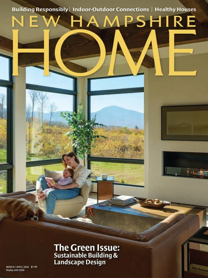 A magazine cover featuring a bright living room with large windows showing a scenic outdoor landscape with trees and mountains. Inside, a woman and a child are sitting on a white armchair, reading a book, with a dog resting on a leather sofa nearby. The room has a modern fireplace and a coffee table with decorative items. The magazine is titled "New Hampshire Home," focusing on sustainable building and landscape design.