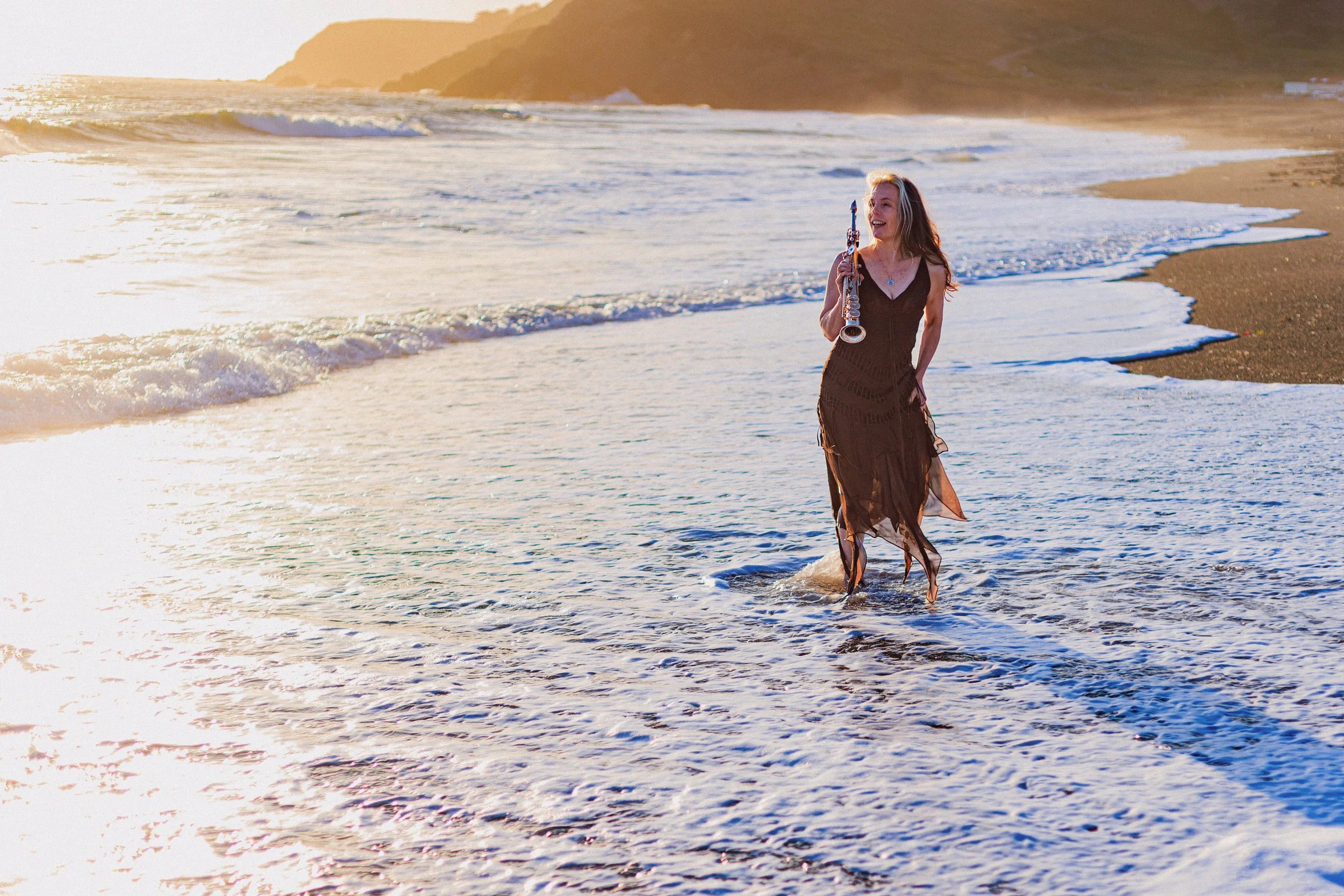 A woman in a black dress walking along the shoreline holding a clarinet at sunset, with waves and hills in the background.