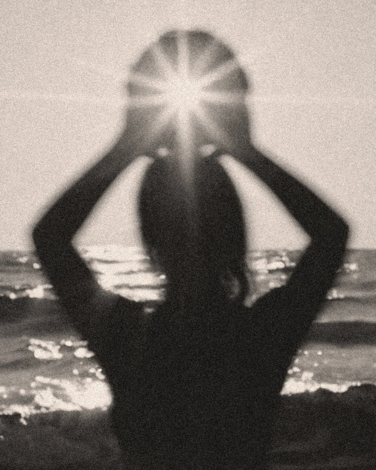 Silhouette of a person with long hair standing by the shoreline at sunset, holding their head with both hands and facing the water.