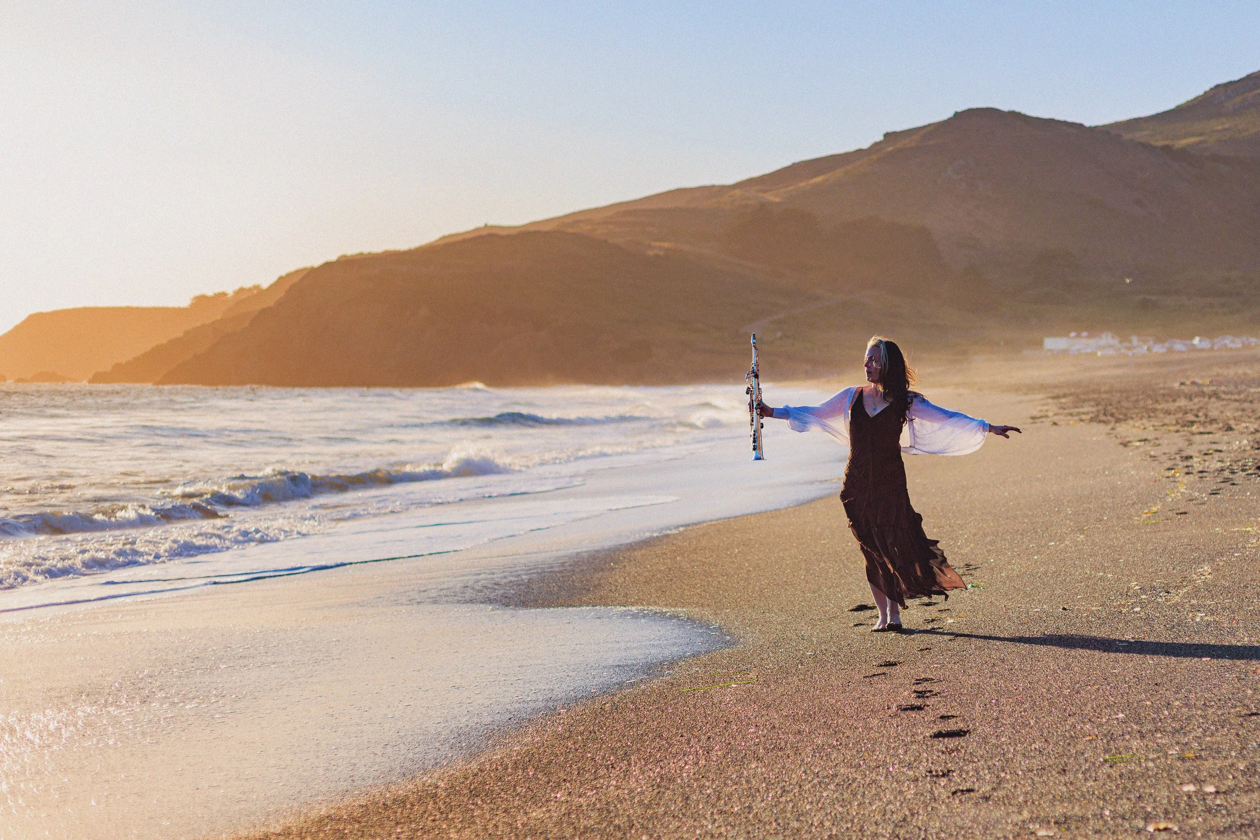 A woman in a long black dress with white sleeves stands on the beach at sunset, holding a musical instrument and spreading her arms with the ocean and mountains in the background.