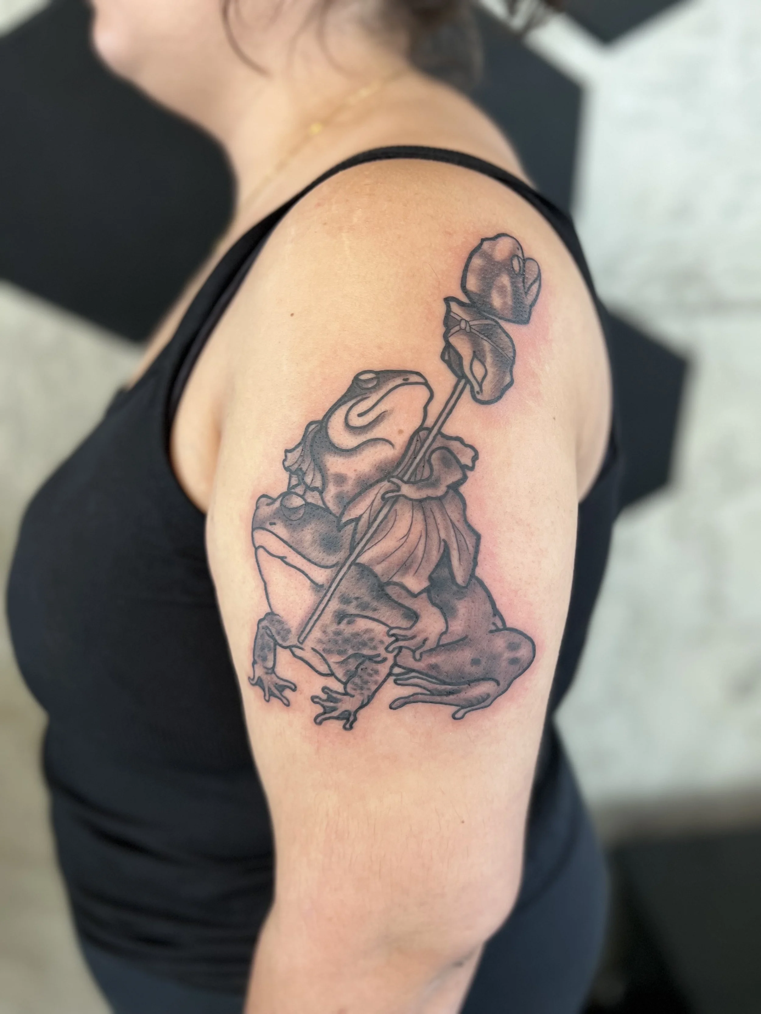 Tattoo of a frog sitting on a lily pad and holding a flower on a person's upper arm.