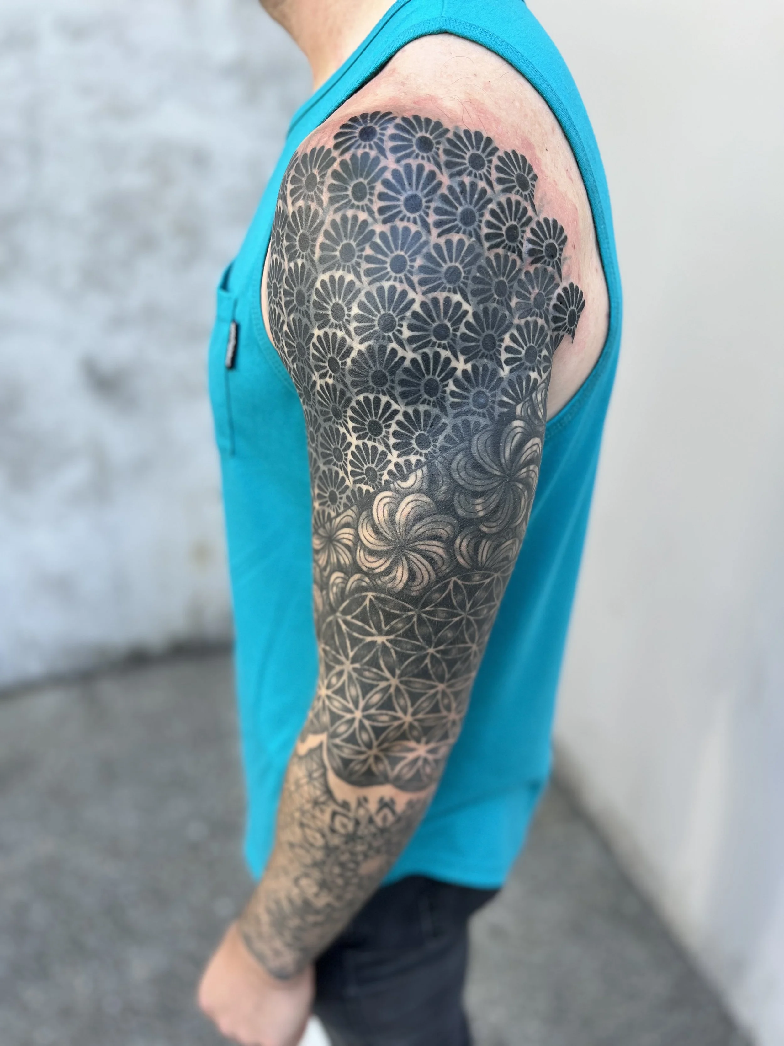 Close-up of a person's upper arm with a tattoo sleeve featuring floral and geometric patterns, wearing a sleeveless turquoise shirt against a gray background.