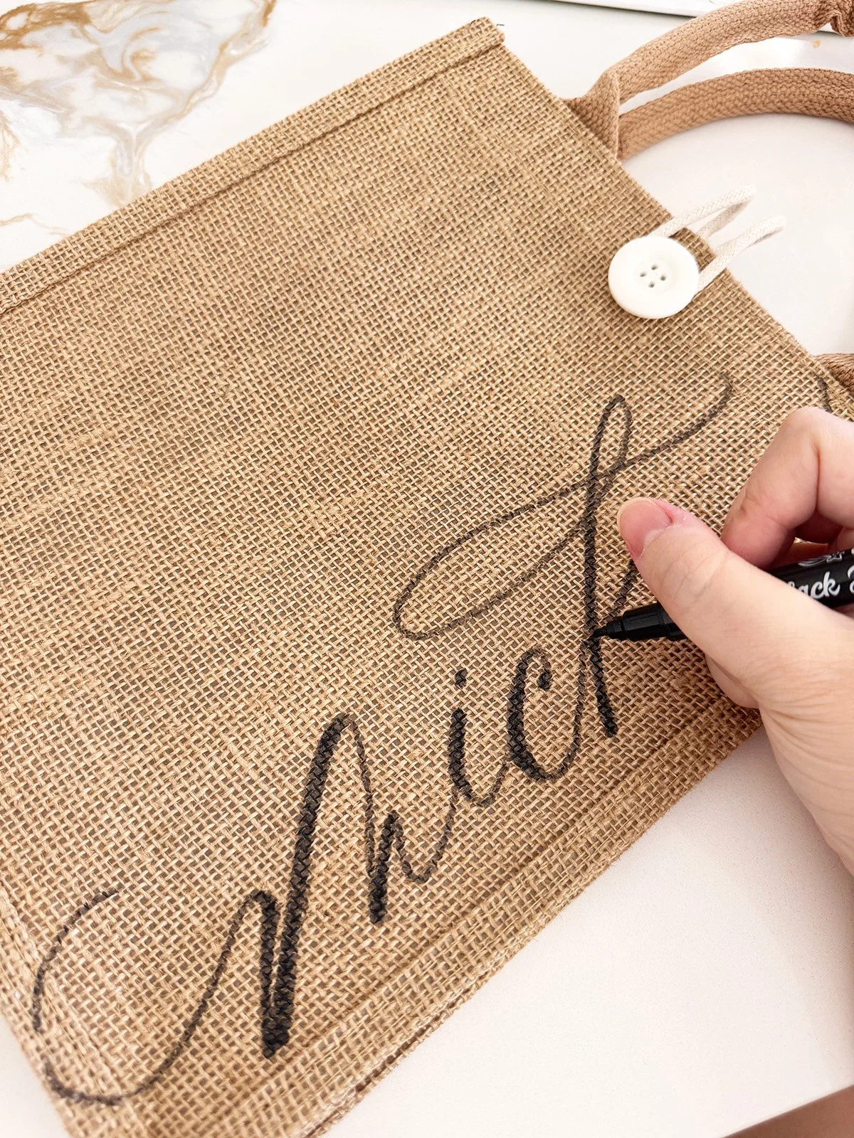 A person is writing a signature with a black marker on a brown, burlap-style tote bag that has the word 'Cheers' written on it in black ink. The tote bag has a white button and a beige strap, resting on a white surface.