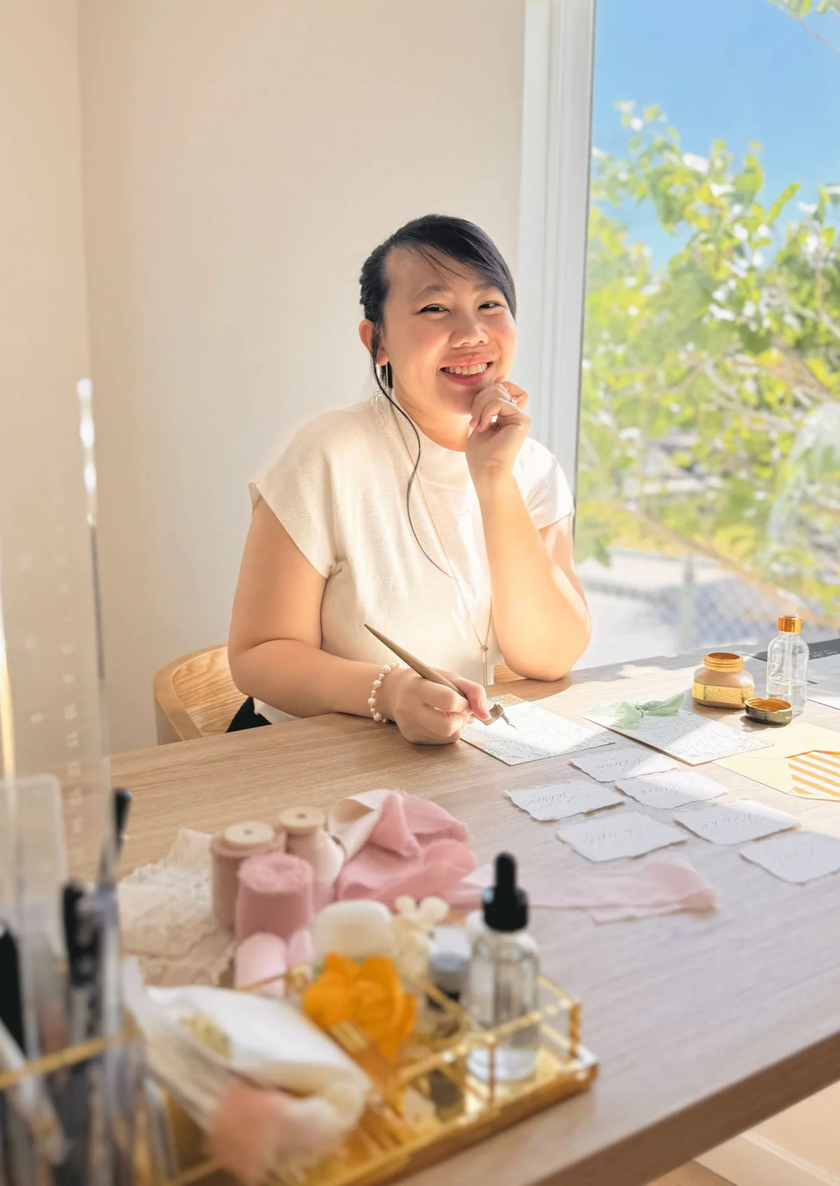 A woman sitting at a wooden table with art supplies, smiling and holding a paintbrush. The table has small bottles, jars, and pieces of paper, with a large window showing green trees and a blue sky outside.
