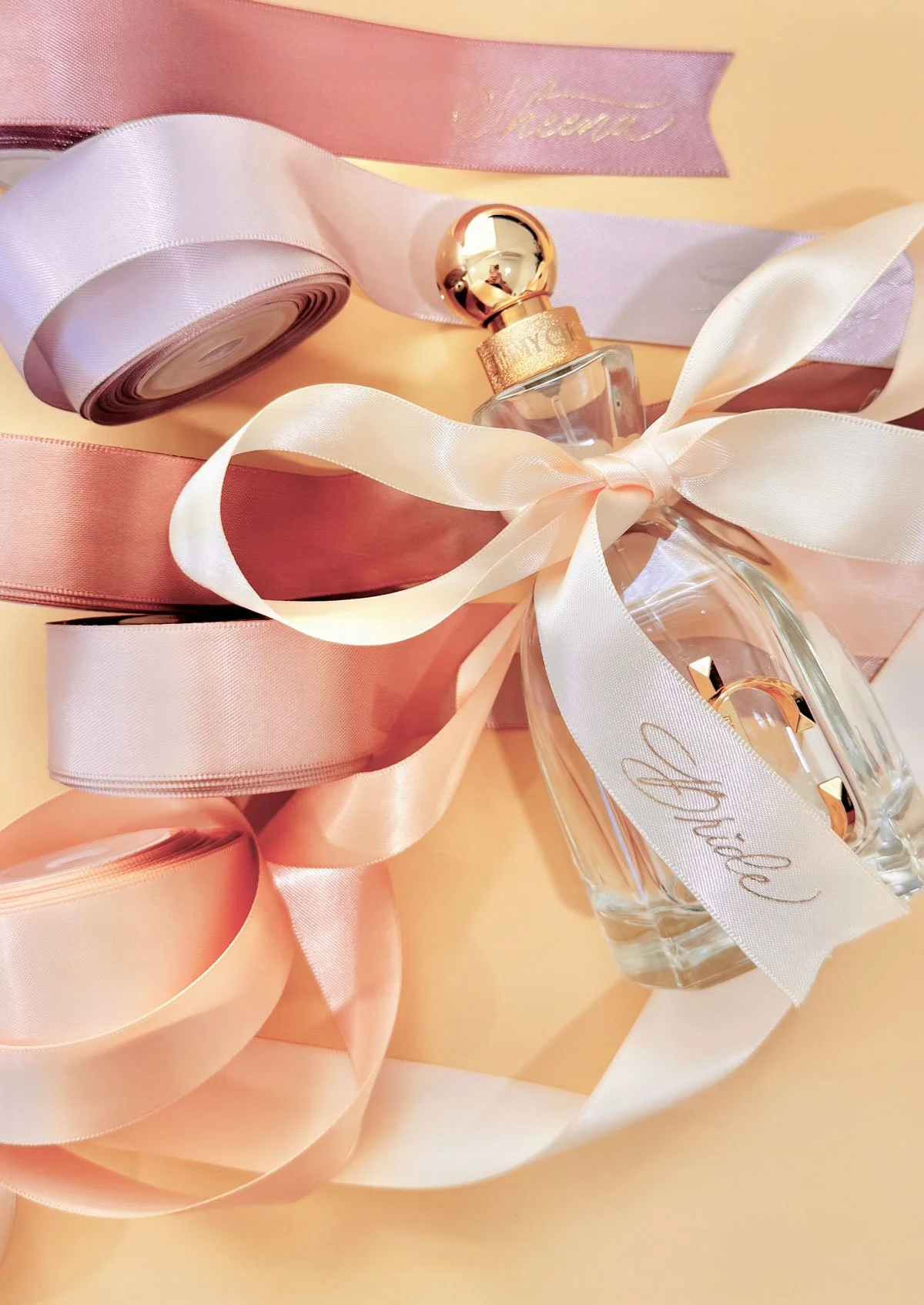 A clear perfume bottle with a gold cap, tied with a white ribbon that has 'Bride' written on it, accompanied by pink and peach ribbons and a pink box with gold writing.