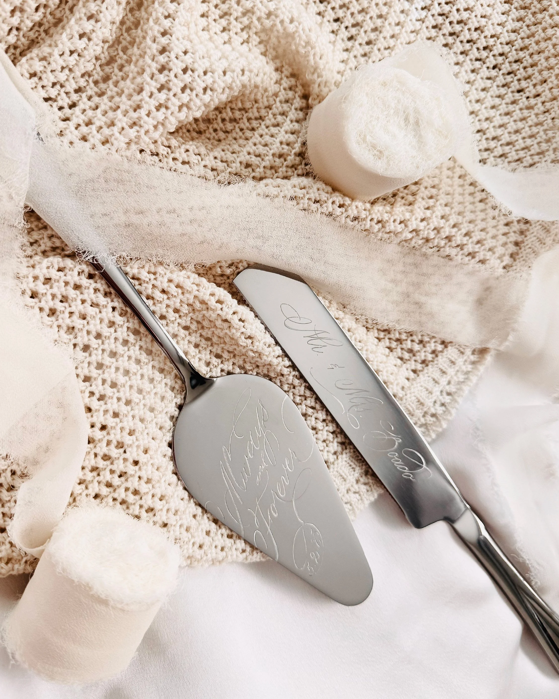 Close-up of a cake knife and server with engraved lettering, surrounded by light-colored fabric, cotton balls, and a crocheted cloth, on a white surface.
