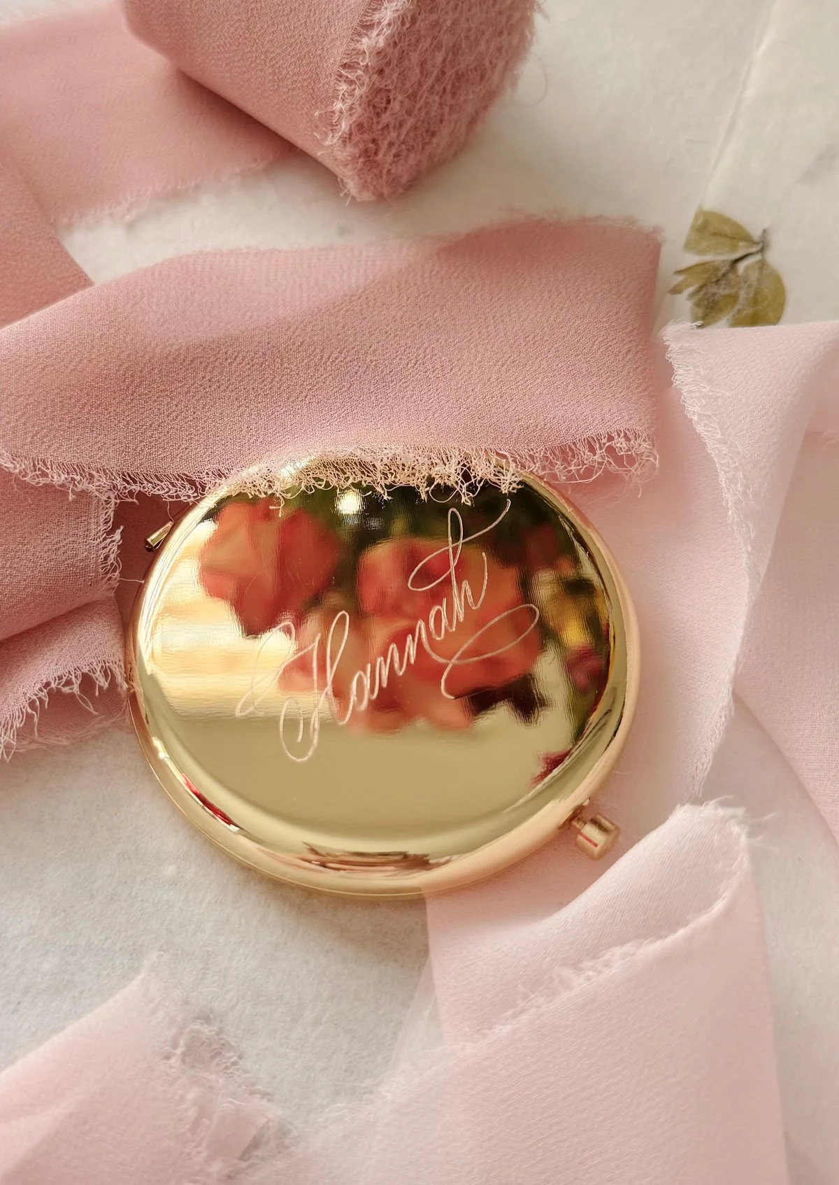 A round, gold-colored compact mirror with the name 'Hannah' engraved in cursive, surrounded by pink fabric and soft pink tissue paper.