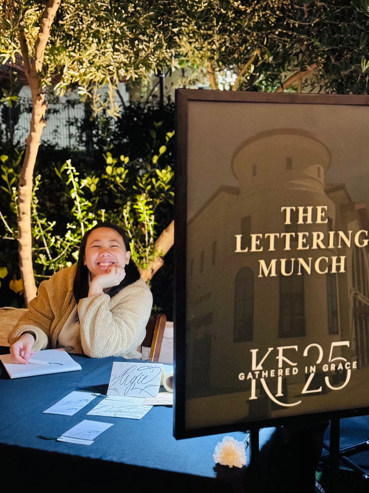A smiling woman with long dark hair wearing a beige coat sitting at a table outdoors at night with trees in the background. The table has papers, a pen, and a name card that says 'Maggie.' To her right, a banner reads 'The Lettering Munch' with additional text beneath.