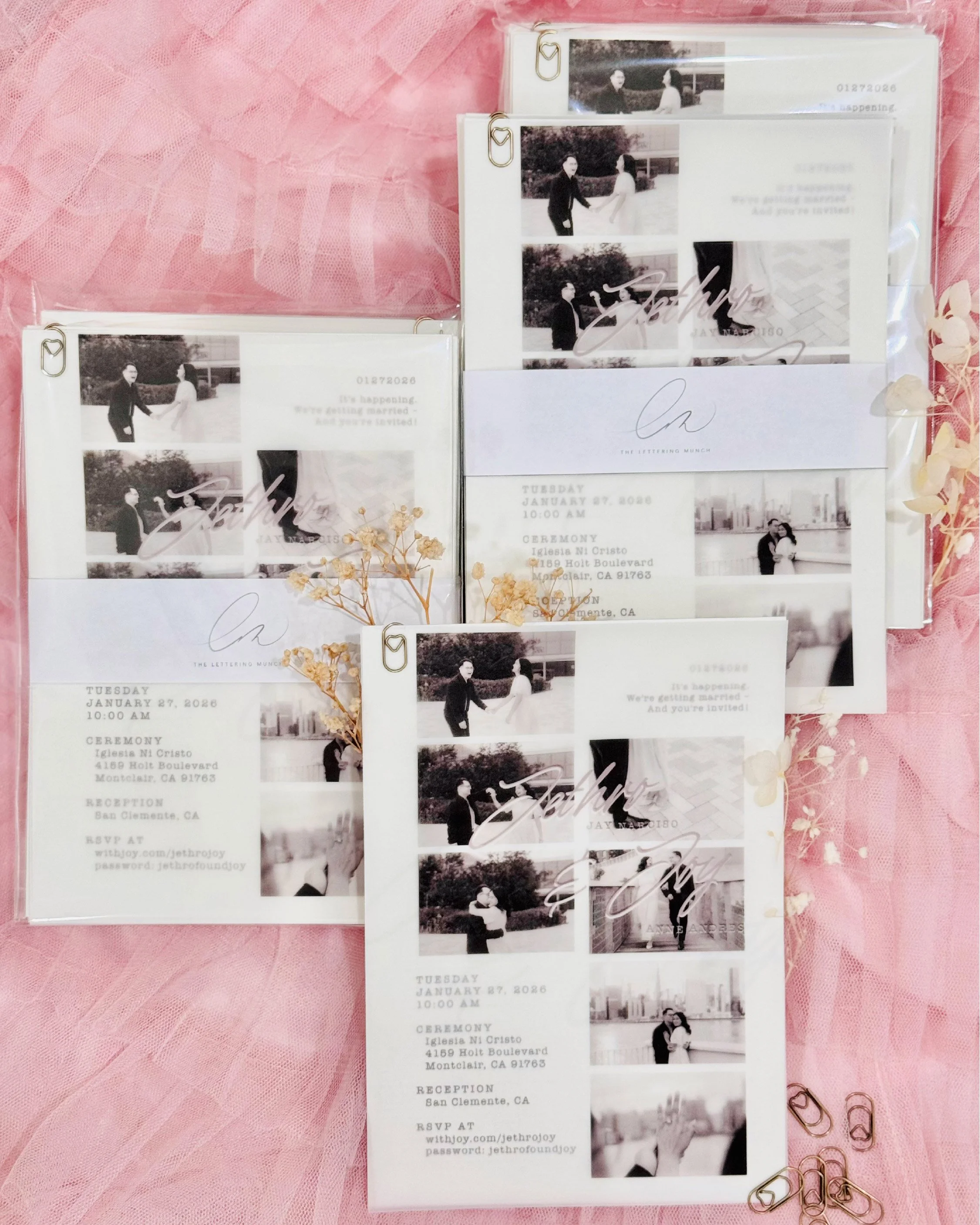 A wedding invitation with black and white photos of a couple holding hands, signed with a cursive signature, and gold paperclips attached to the edges, placed on a pink textured surface with pink paperclips nearby.
