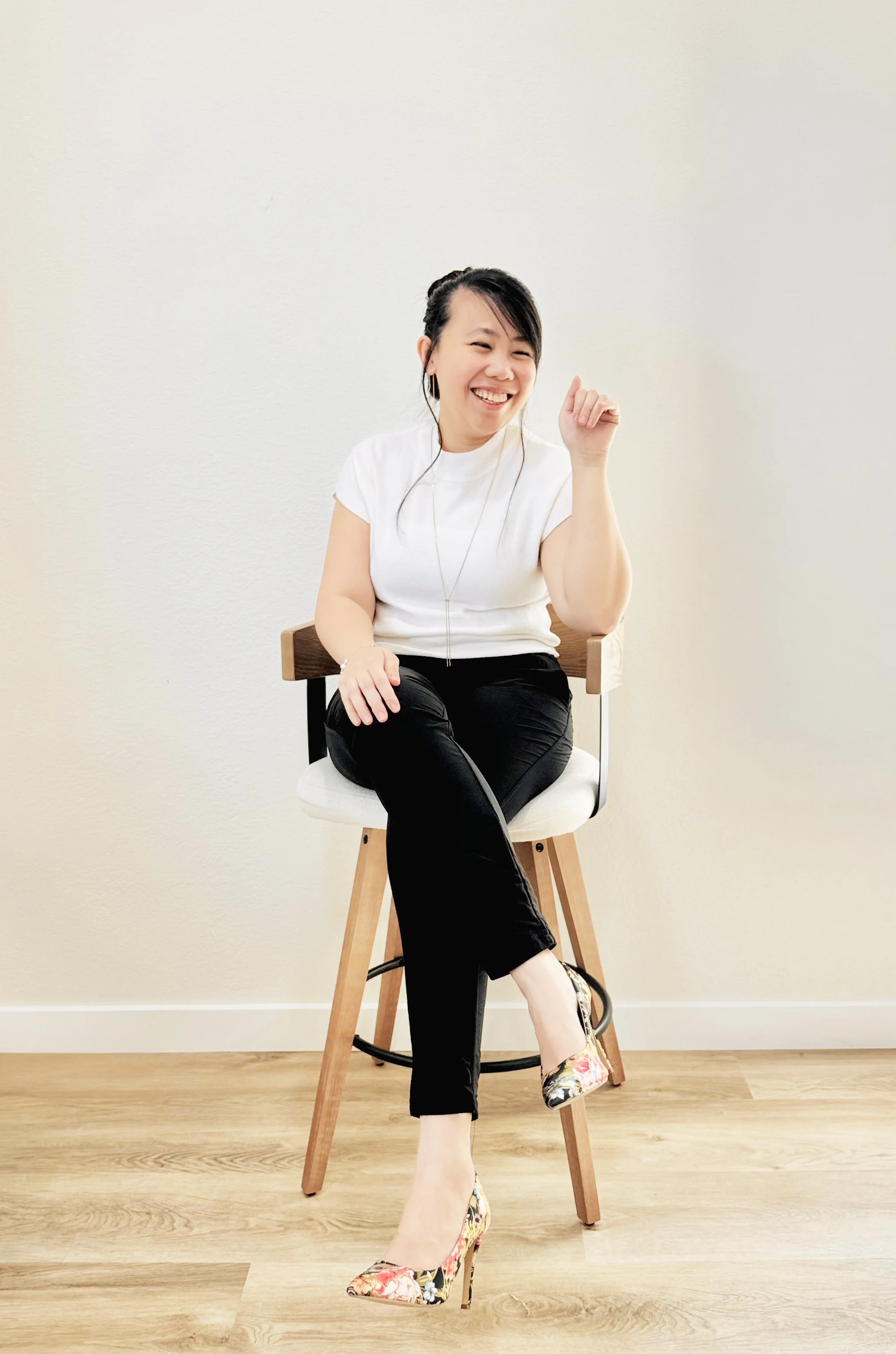 A woman with black hair, wearing a white t-shirt and black pants, sitting on a high stool with wooden legs, smiling and raising her right fist, in a room with beige walls and wooden floor, wearing floral high heels.