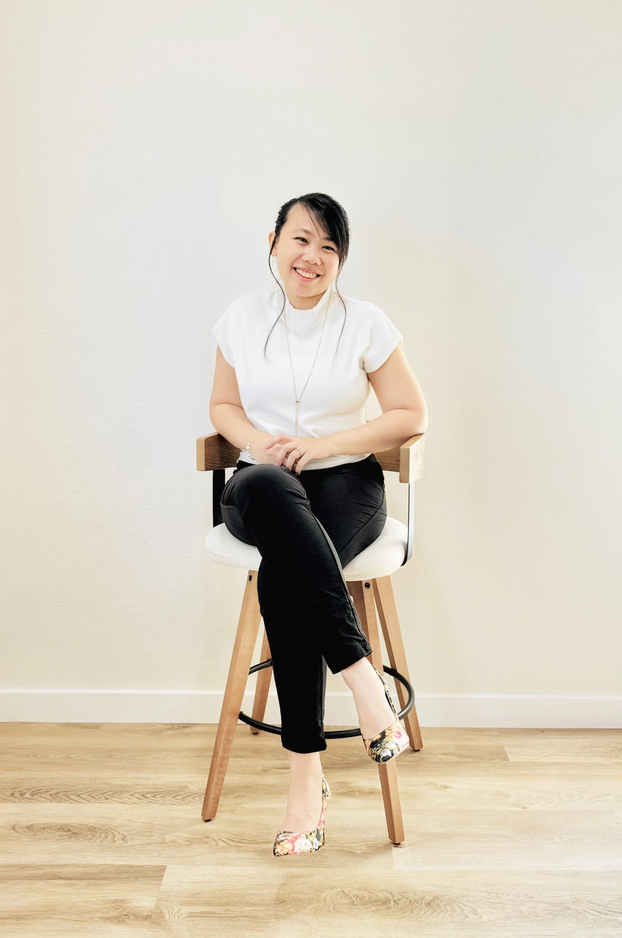 A woman sitting on a wooden chair with a white cushion, smiling, against a plain off-white wall, wearing a white top, black pants, and floral high heels.