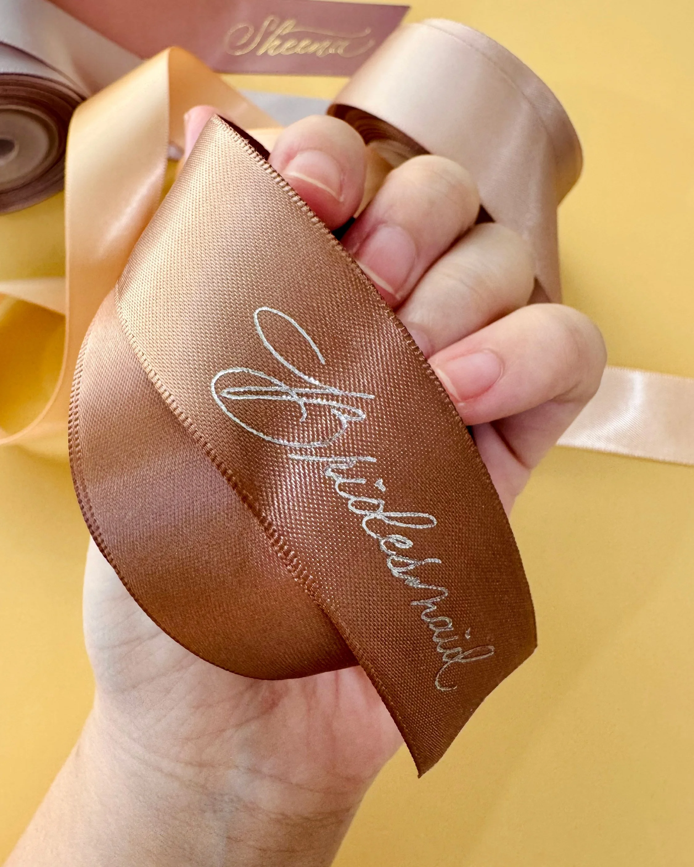 Close-up of a hand holding a shiny bronze-colored ribbon with white cursive writing, against a yellow background.